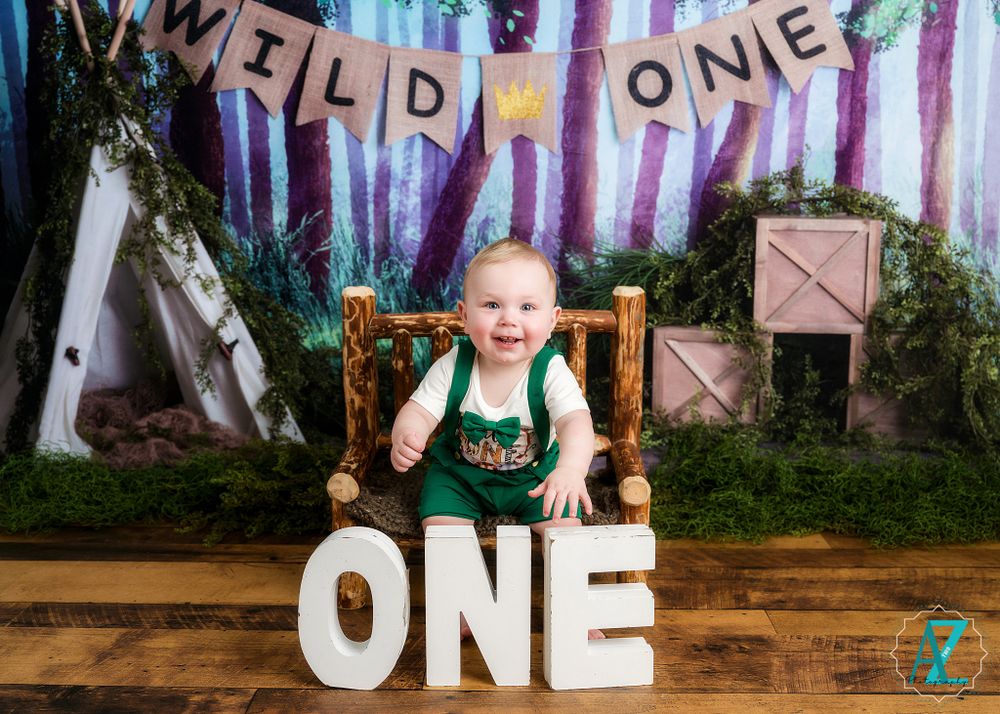 Happy child in green overalls sitting on wooden chair with forest-themed first birthday setup.