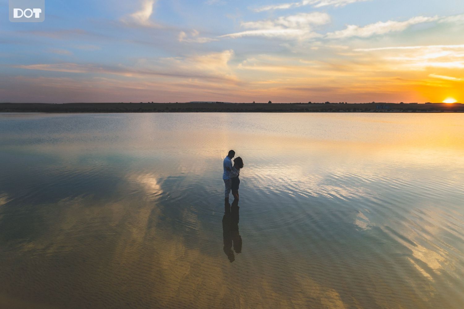 Silhouetted couple reflected in calm water during golden sunset.