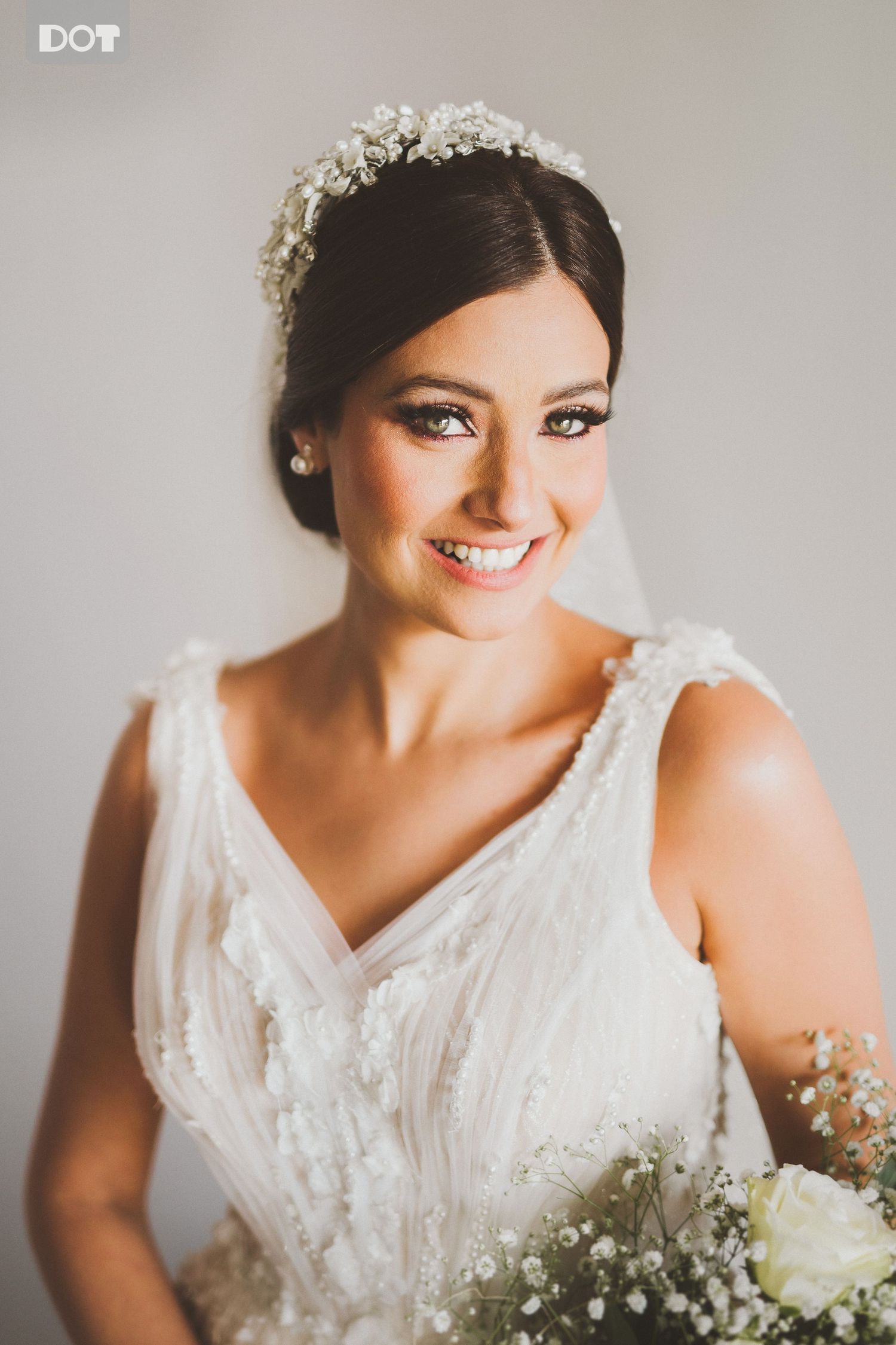 Bride in white lace dress smiles while holding delicate baby's breath bouquet against light background.