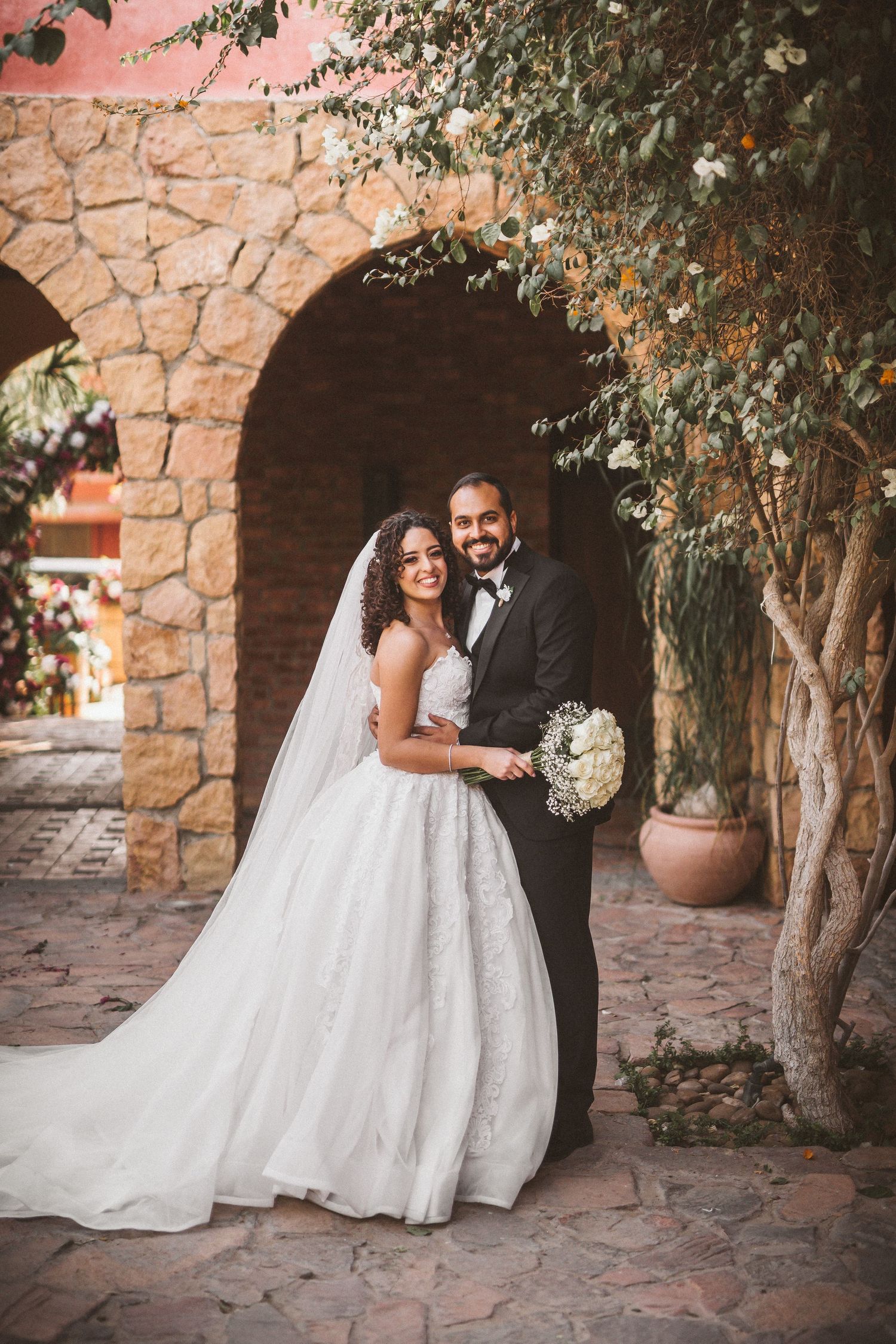 Romantic wedding couple poses against rustic stone wall backdrop with flowing white dress and natural greenery.