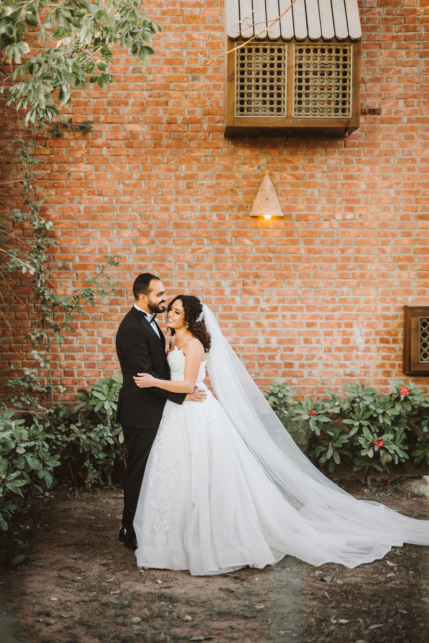 Elegant wedding couple shares intimate moment against red brick wall with trailing vines.