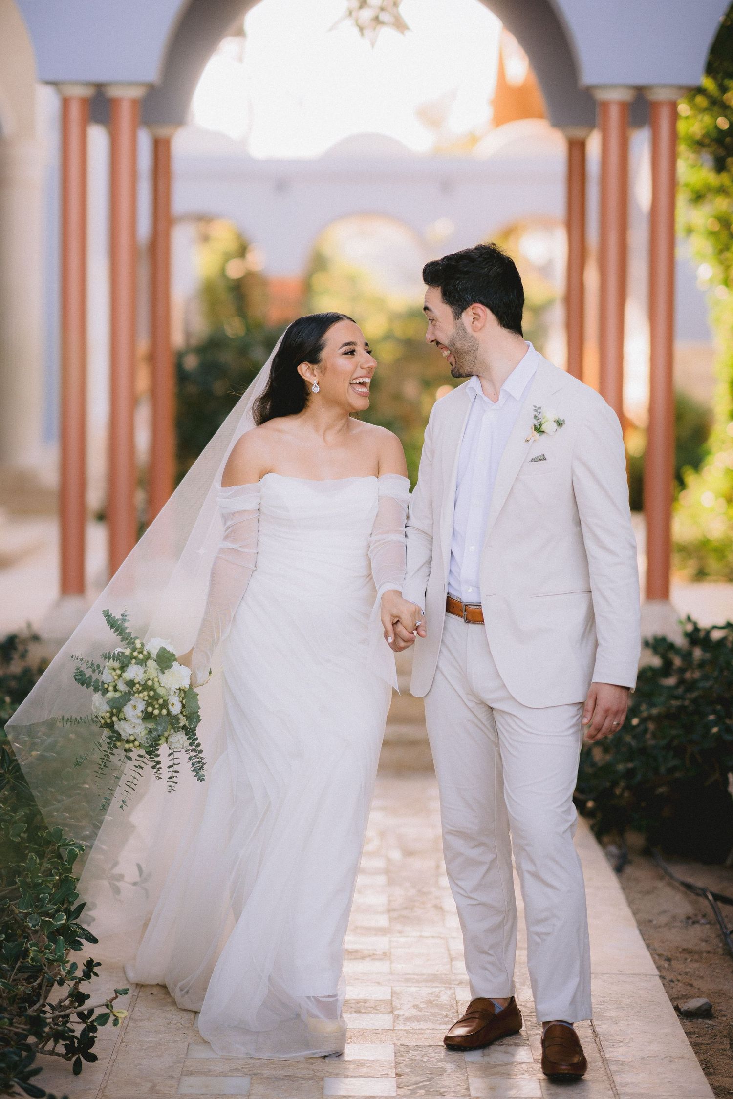Couple walking hand in hand under arched walkway in elegant white wedding attire.