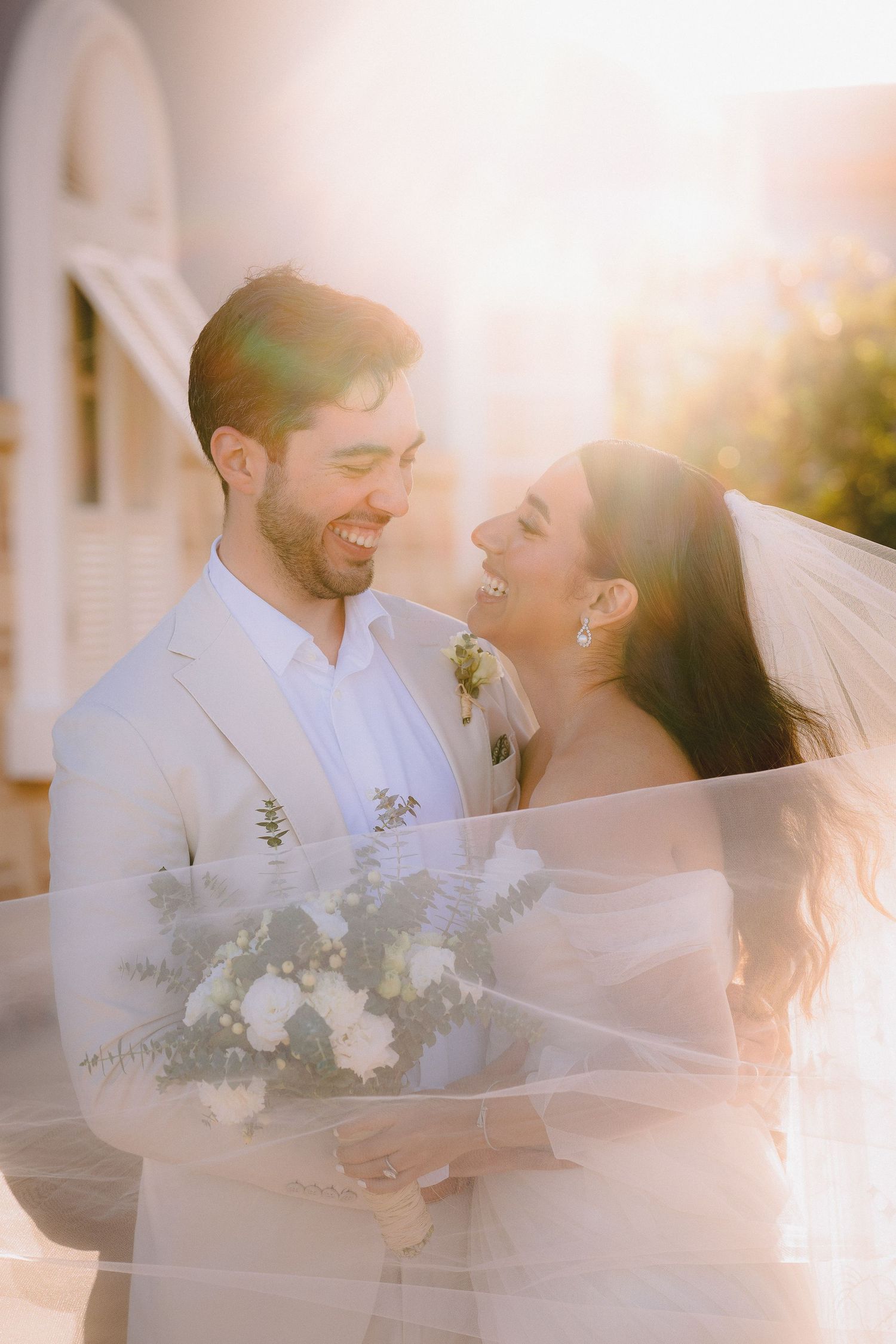 Newlyweds share a joyful moment in golden sunlight while holding a white floral bouquet.