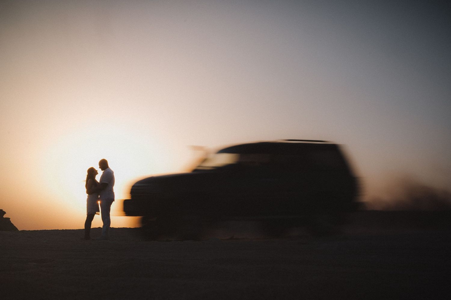 Silhouetted couple stands near parked car against dramatic sunset sky.