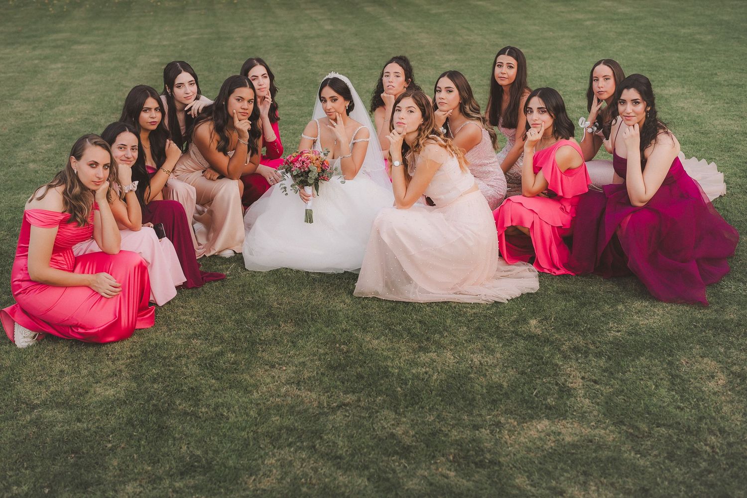 Bridesmaids in pink and coral dresses pose together on a grassy lawn during a wedding photoshoot.