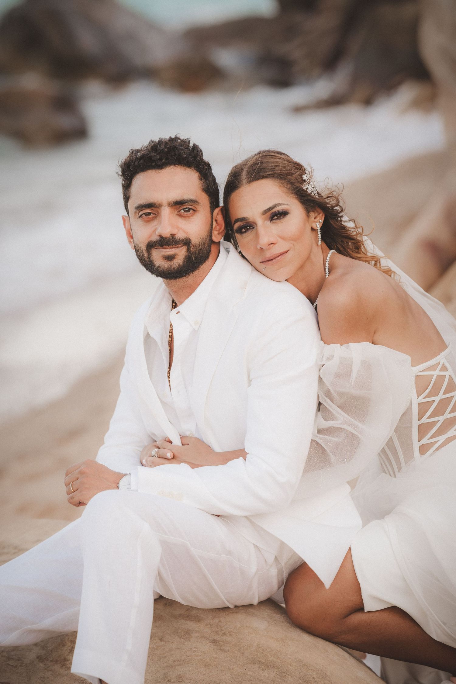 Romantic beach photoshoot with couple in white outfits lounging on sandy shore.
