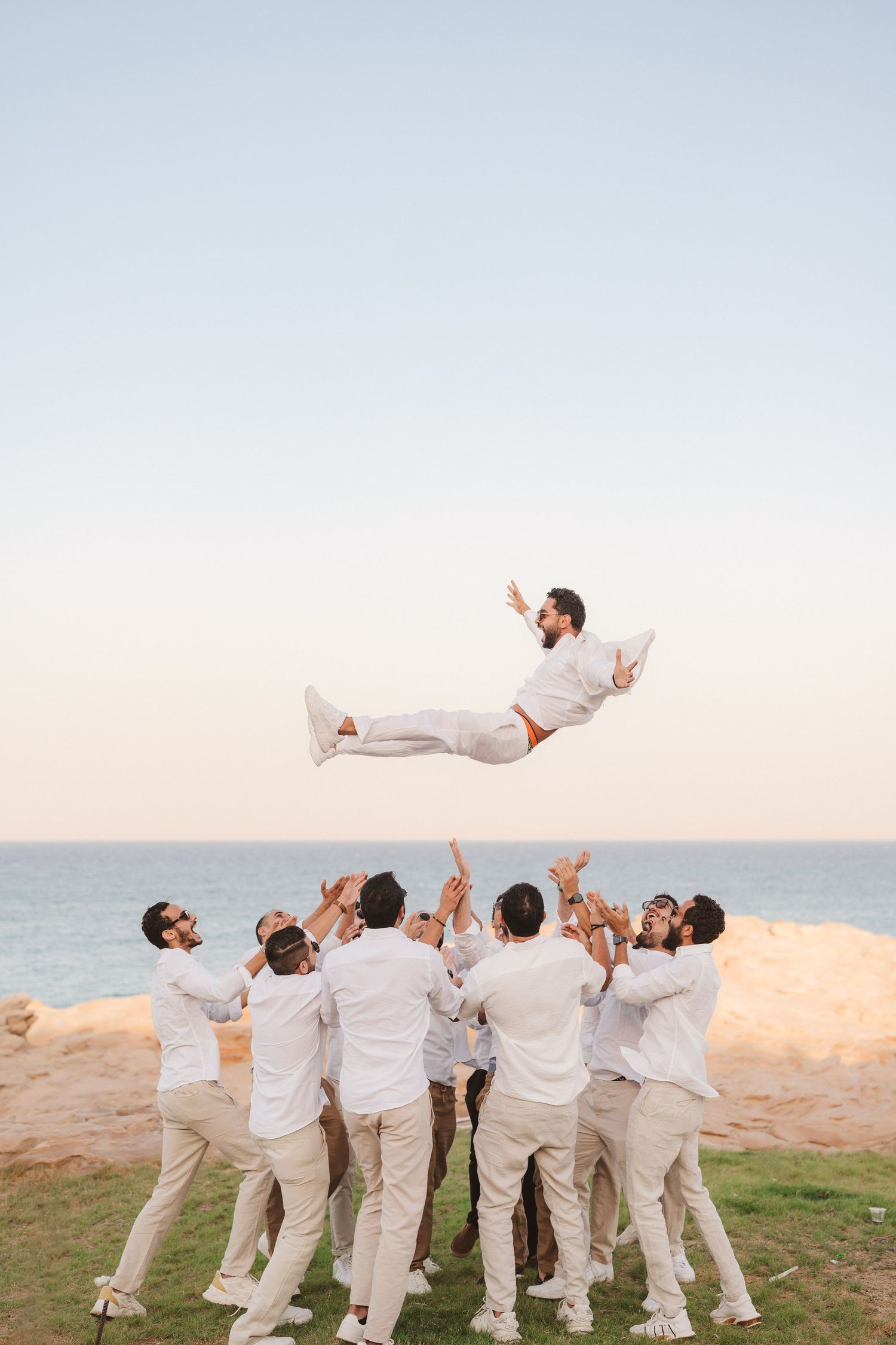 Groomsmen in white attire toss someone into the air at a beach wedding celebration.