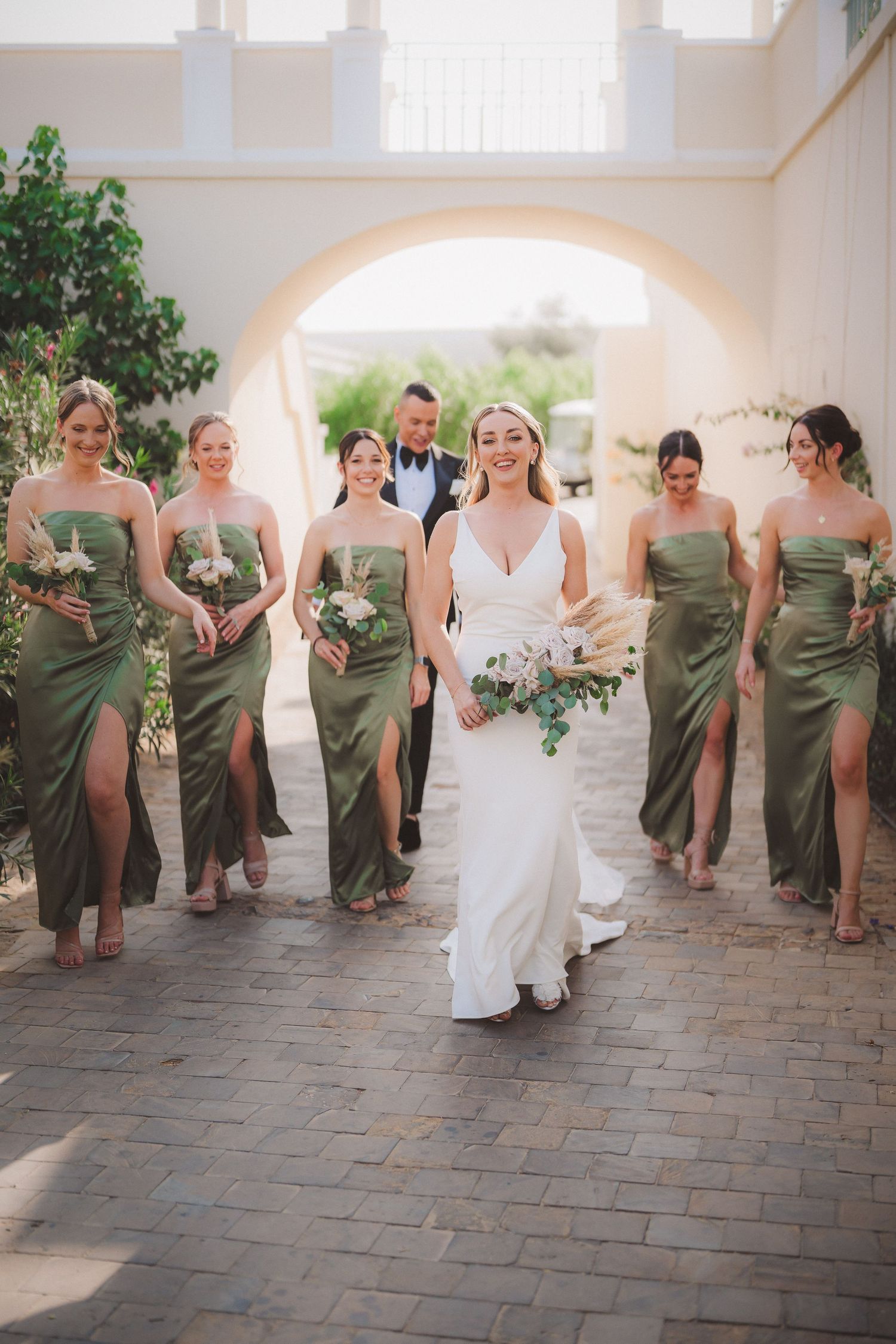 Bridal party in olive green dresses poses playfully in arched hallway.