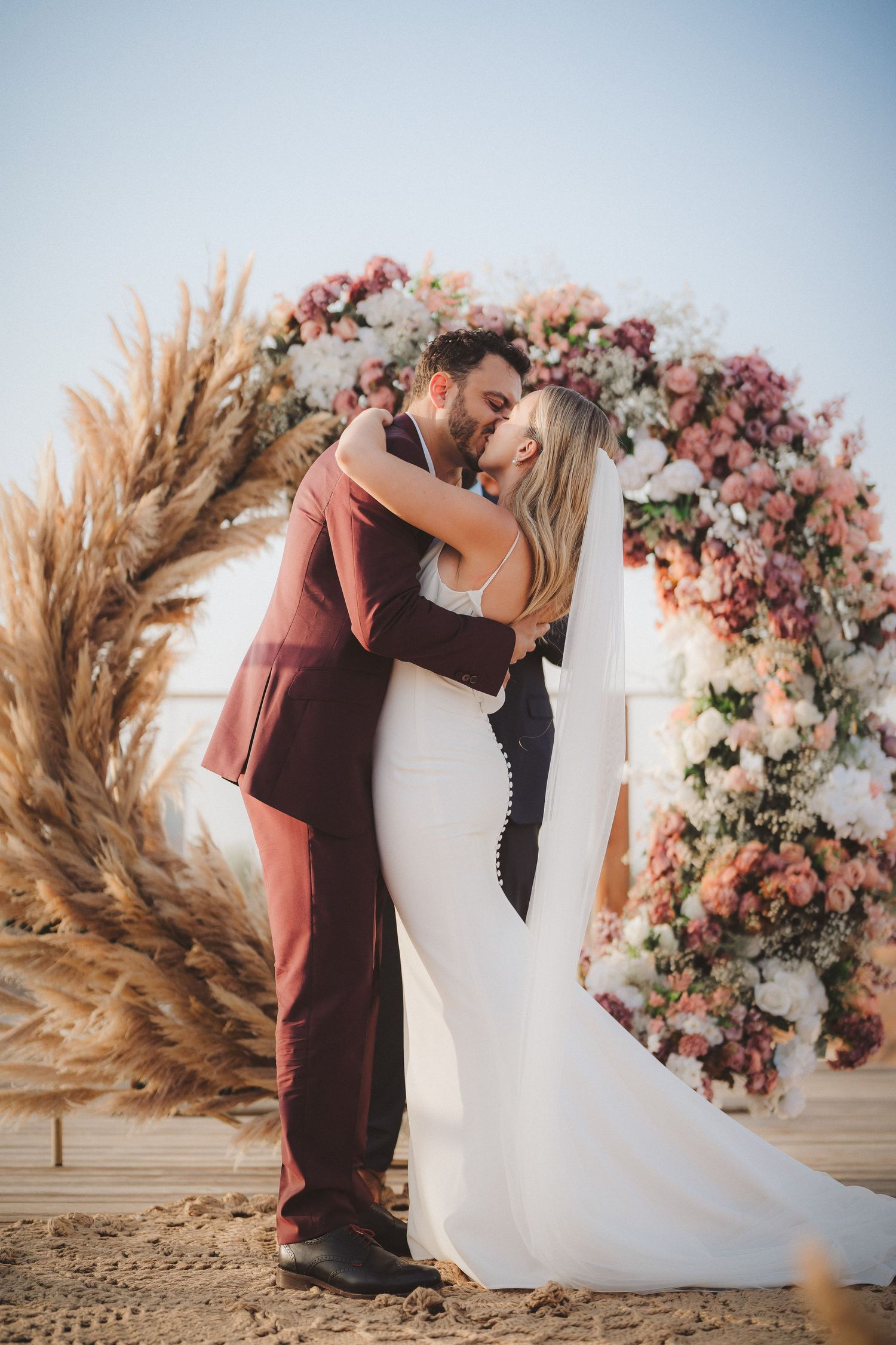 Wedding couple embraces in front of a floral arch with pampas grass and pink roses.