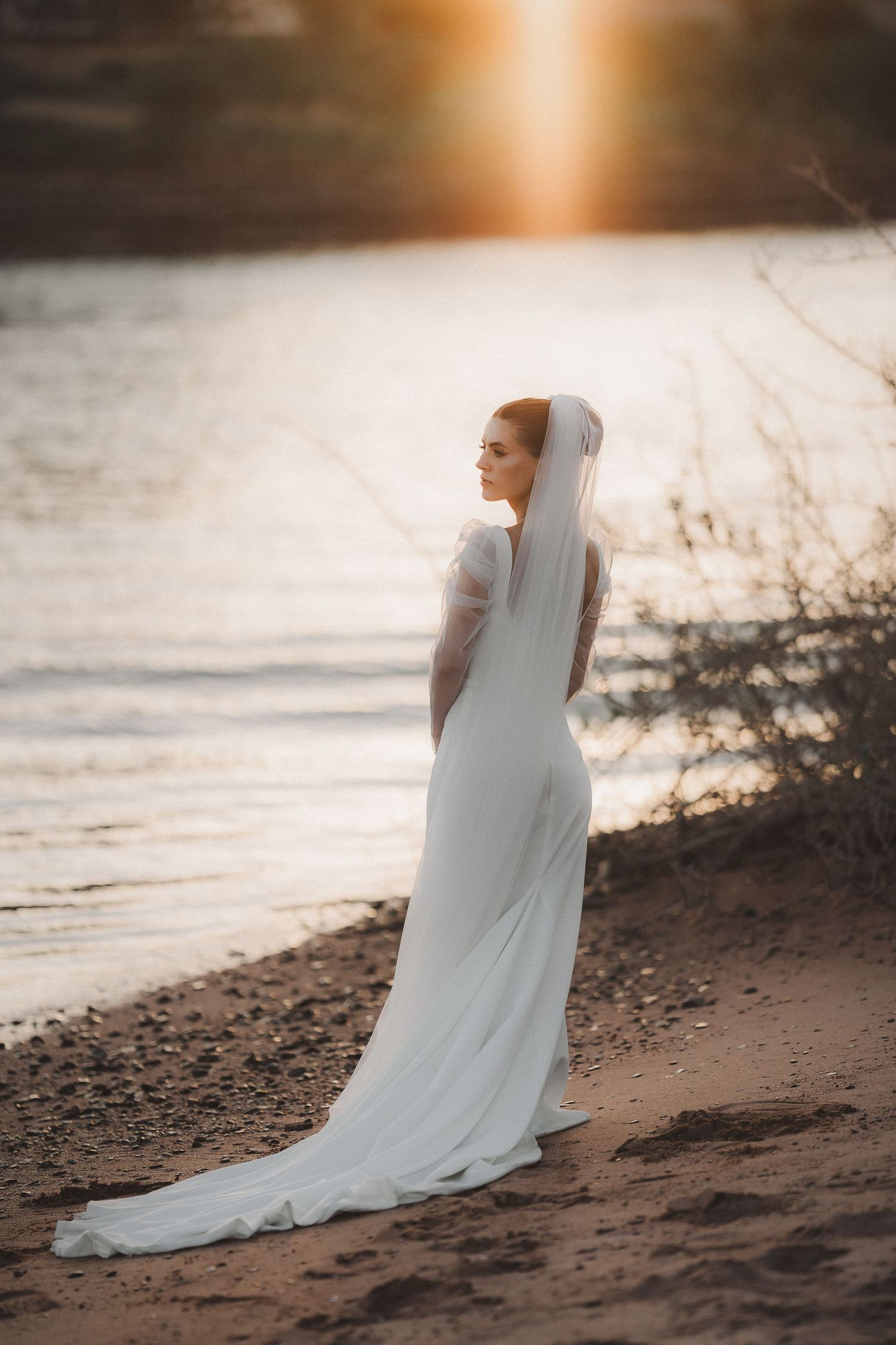 Bride in flowing white dress stands by waterfront during golden sunset.