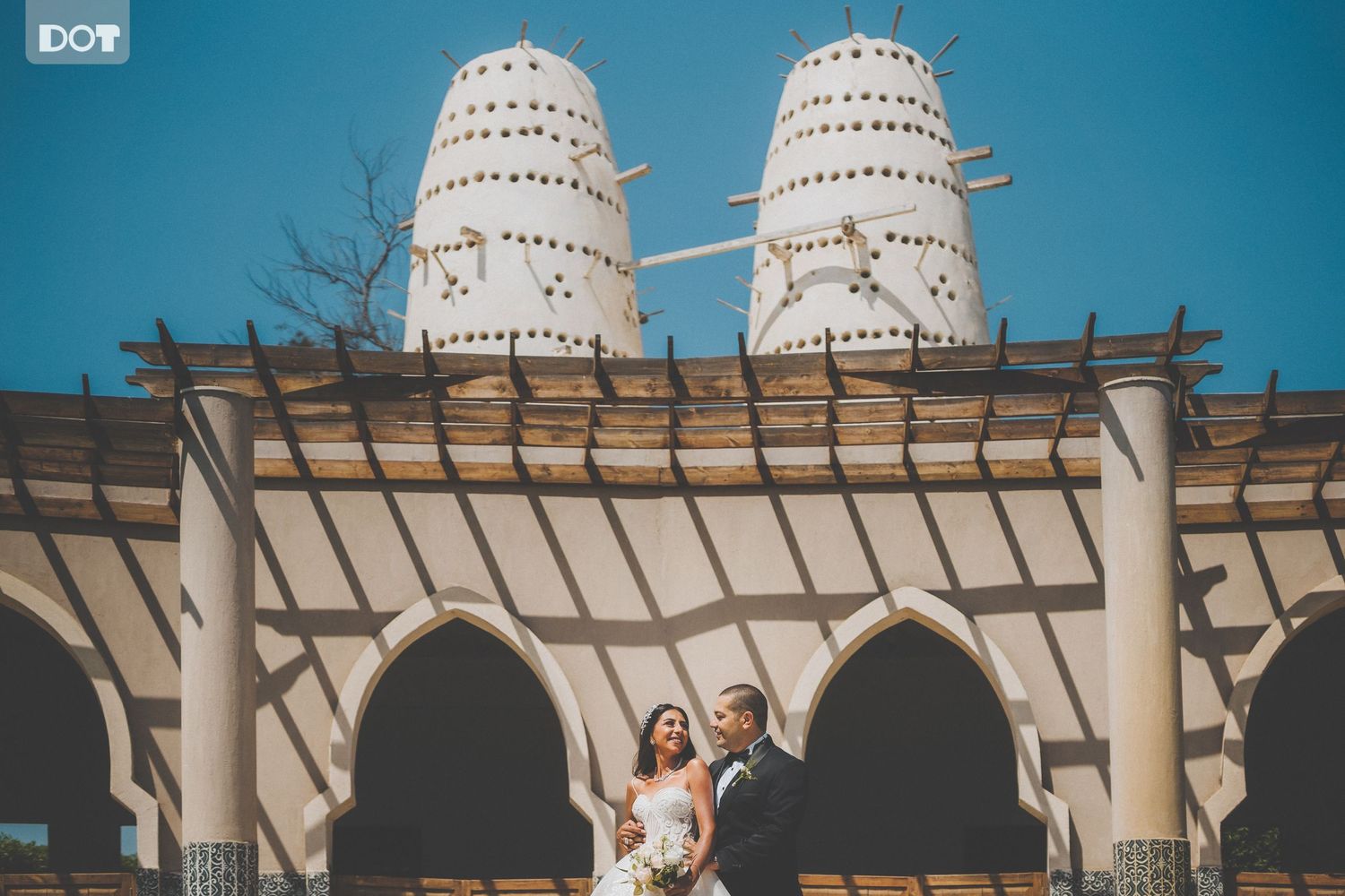 Modern architectural wedding venue with white towers and wooden pergola against blue sky.