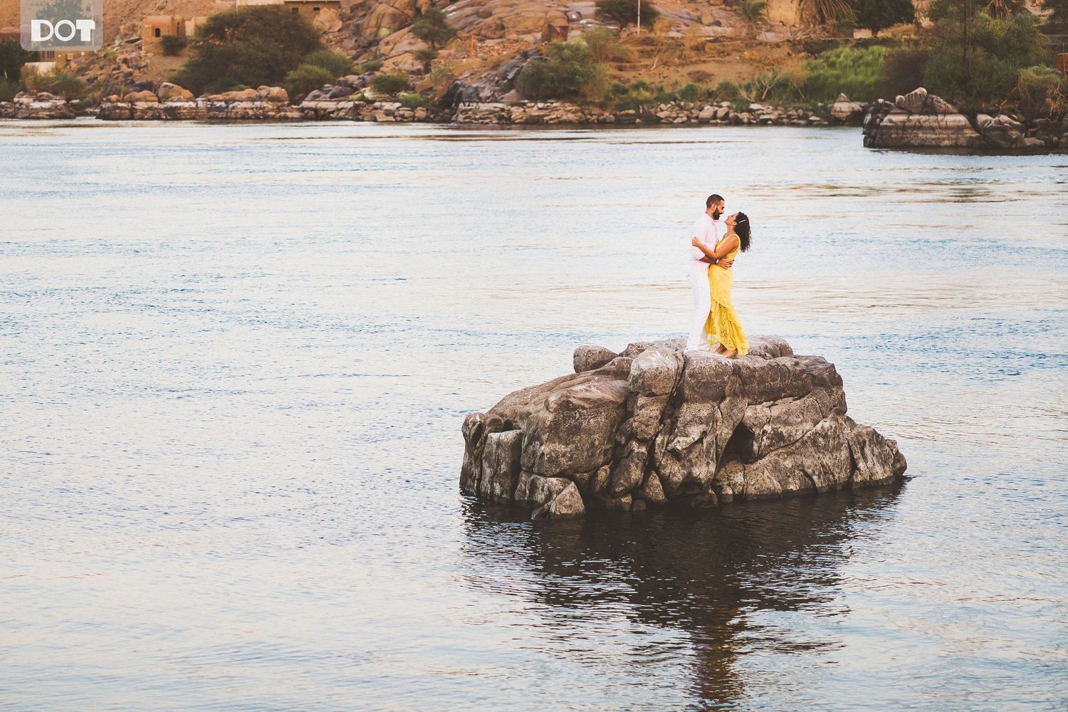 Couple standing on rocky outcrop surrounded by serene ocean waters.