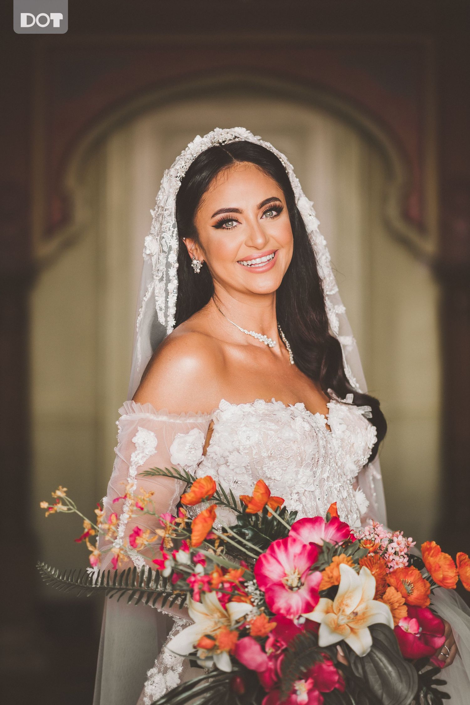 A bride in a lace off-shoulder gown and veil holds a vibrant bouquet of tropical flowers.