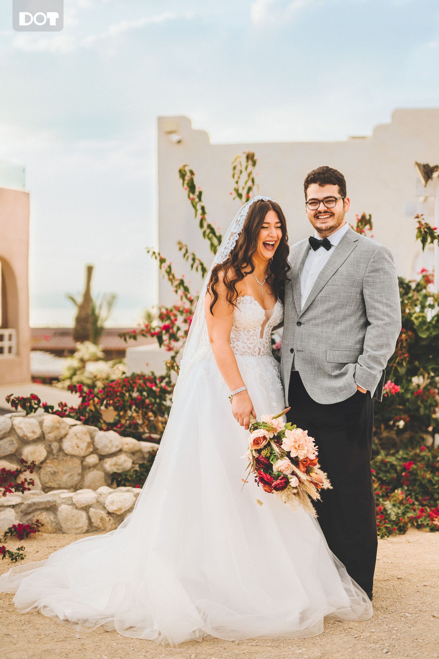 A newlywed couple poses in front of a desert resort with colorful flowers and architecture.