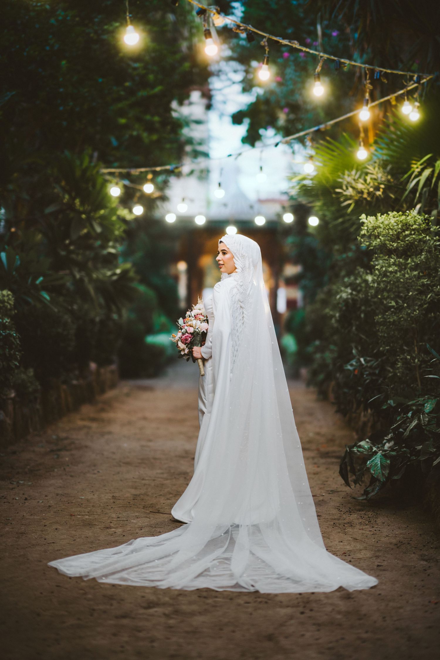 Bride in flowing white gown stands under string lights on garden path.