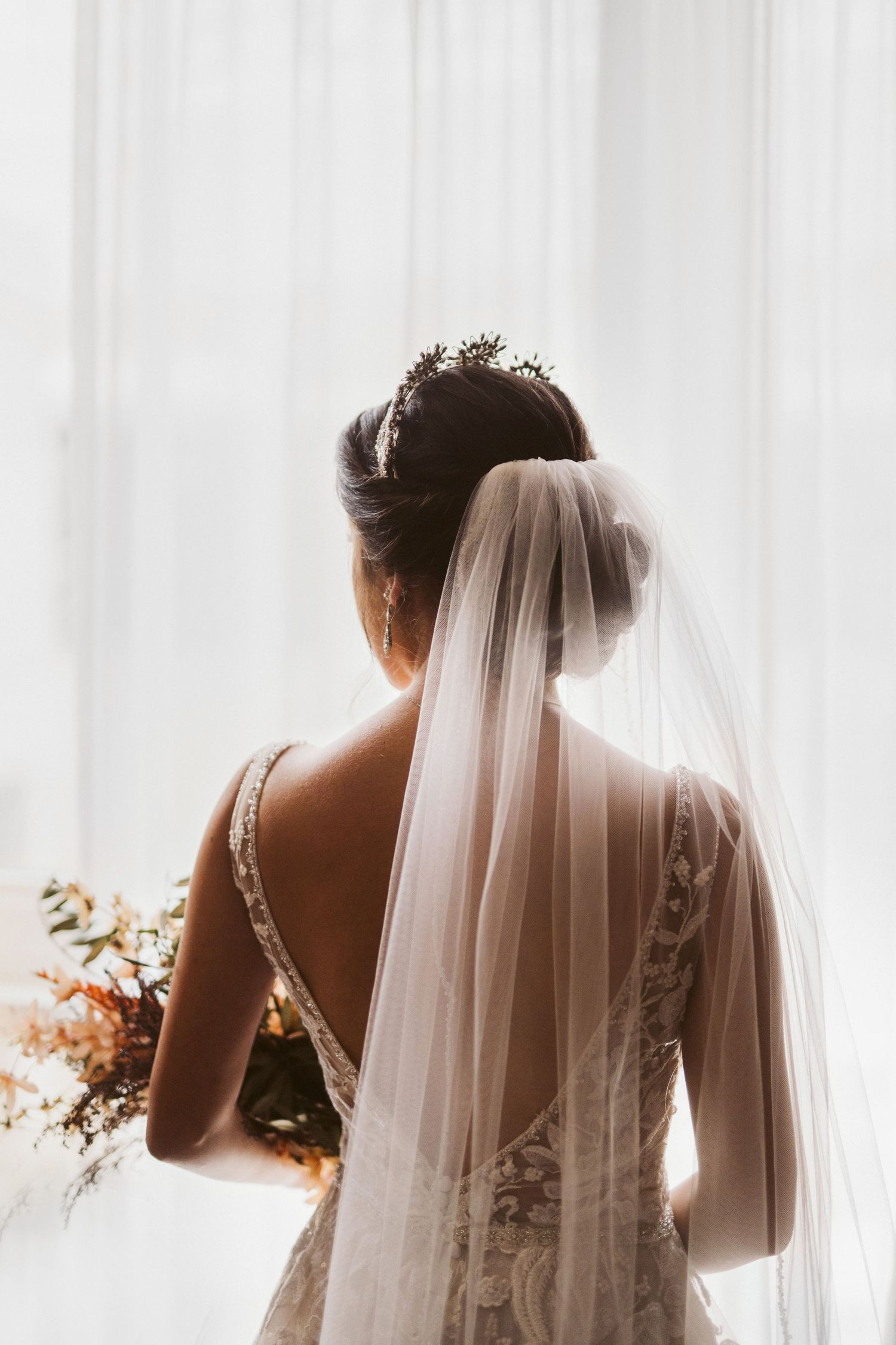 Elegant silhouette of bride getting ready with flowing veil against white curtains.
