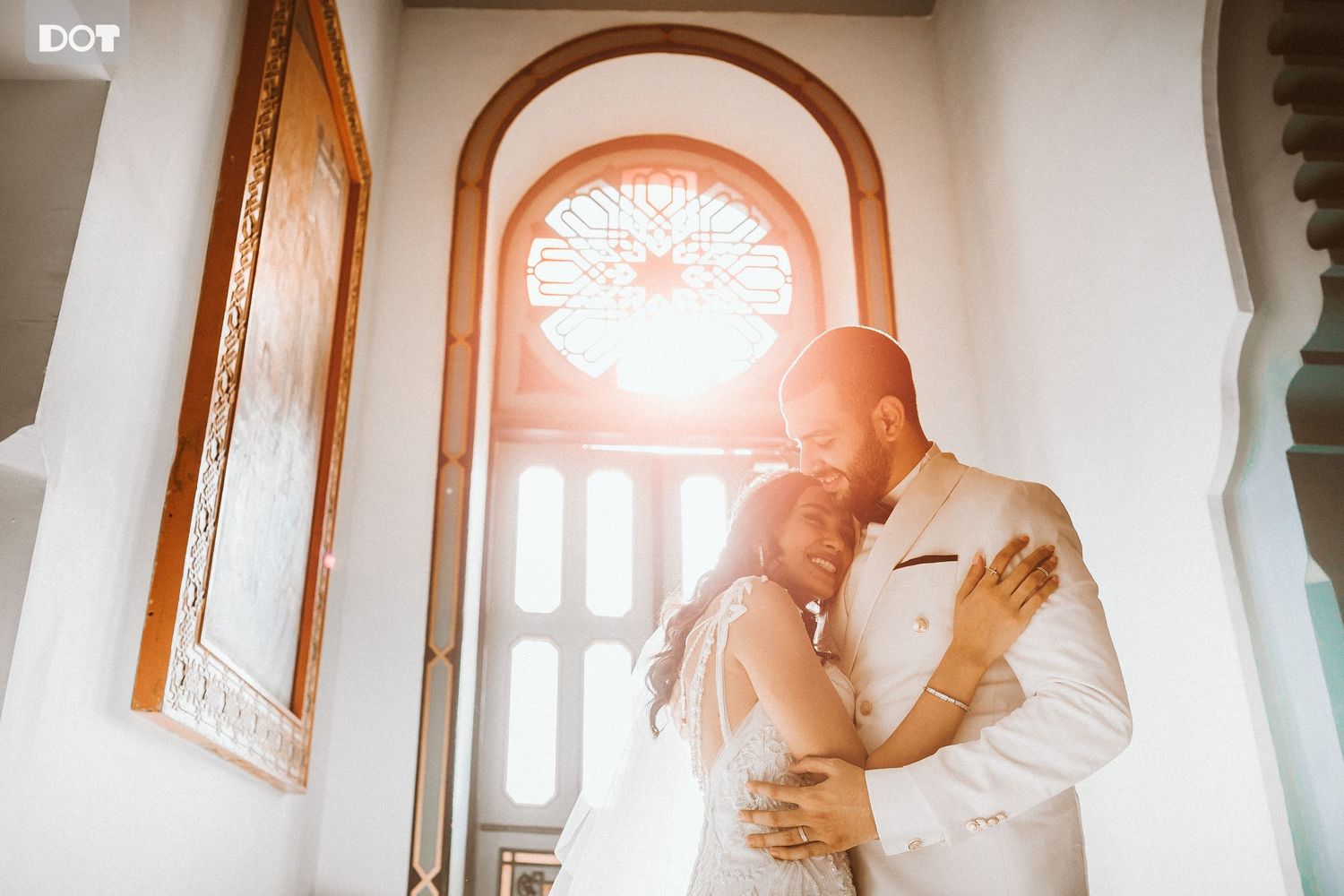 Romantic wedding couple embraces in a sunlit church interior with stained glass windows.