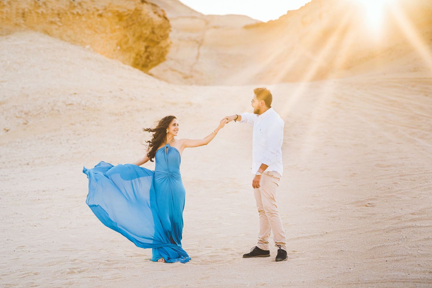 Couple in blue dress and white outfit dances in desert sunlight.