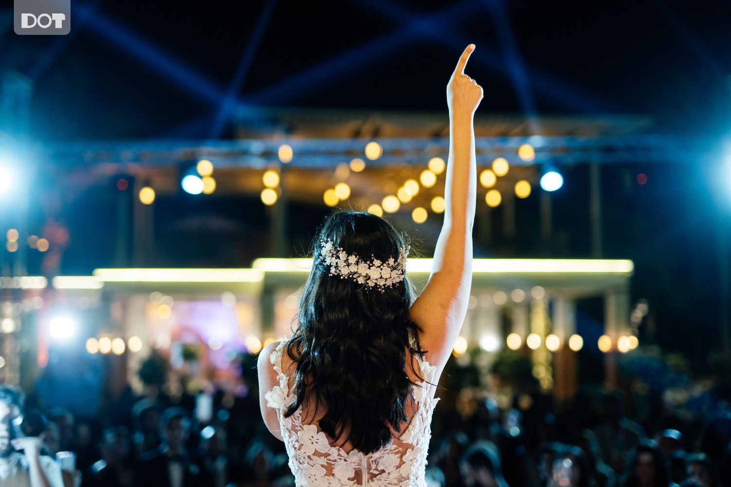 Bride with long dark hair raises arm triumphantly at evening reception with string lights.