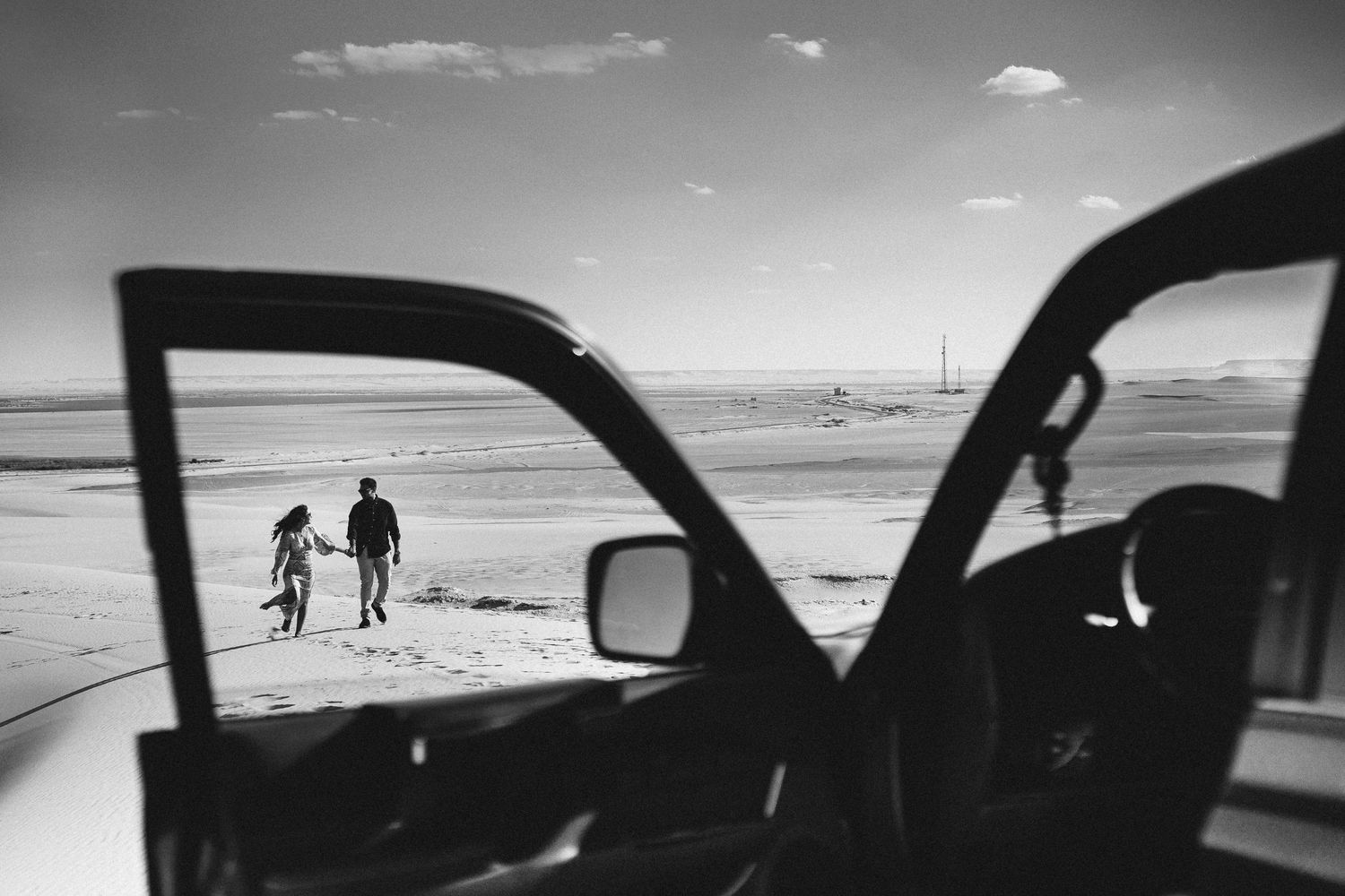 Black and white photo of two figures walking on beach, viewed through car window.