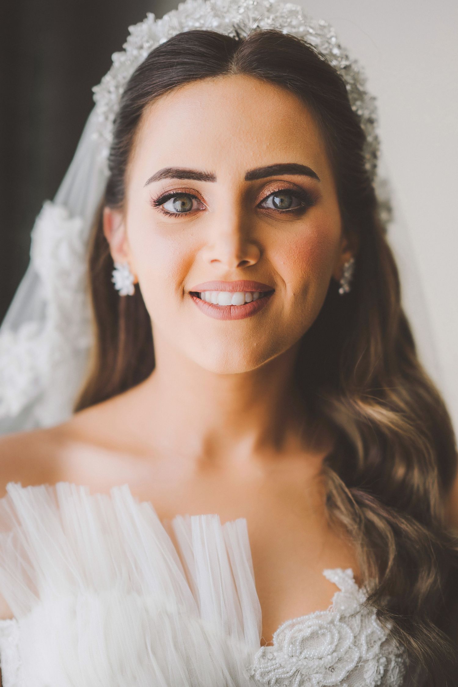 Bride wearing lace veil and crystal headpiece smiles in natural window light.