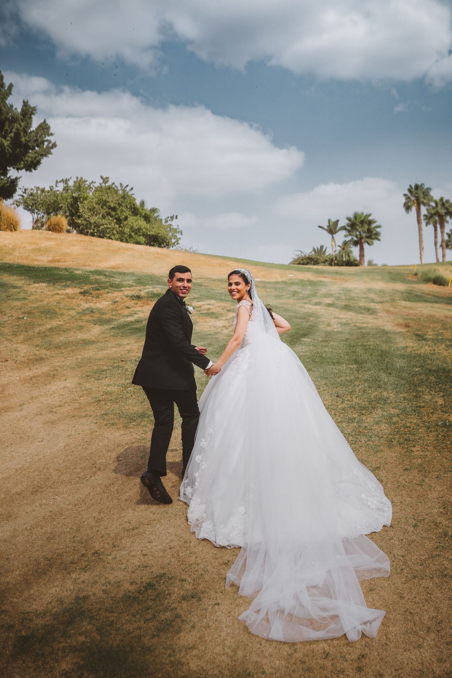 Couple in wedding attire pose on hill path with flowing dress train and palm trees.