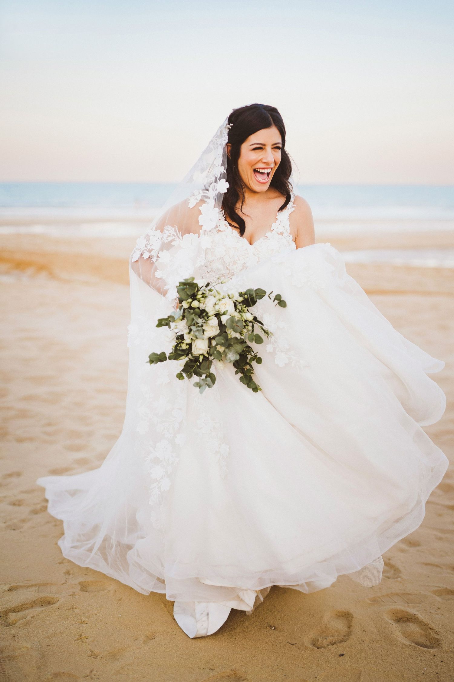 Joyful bride in flowing white dress laughs while holding bouquet on sandy beach.