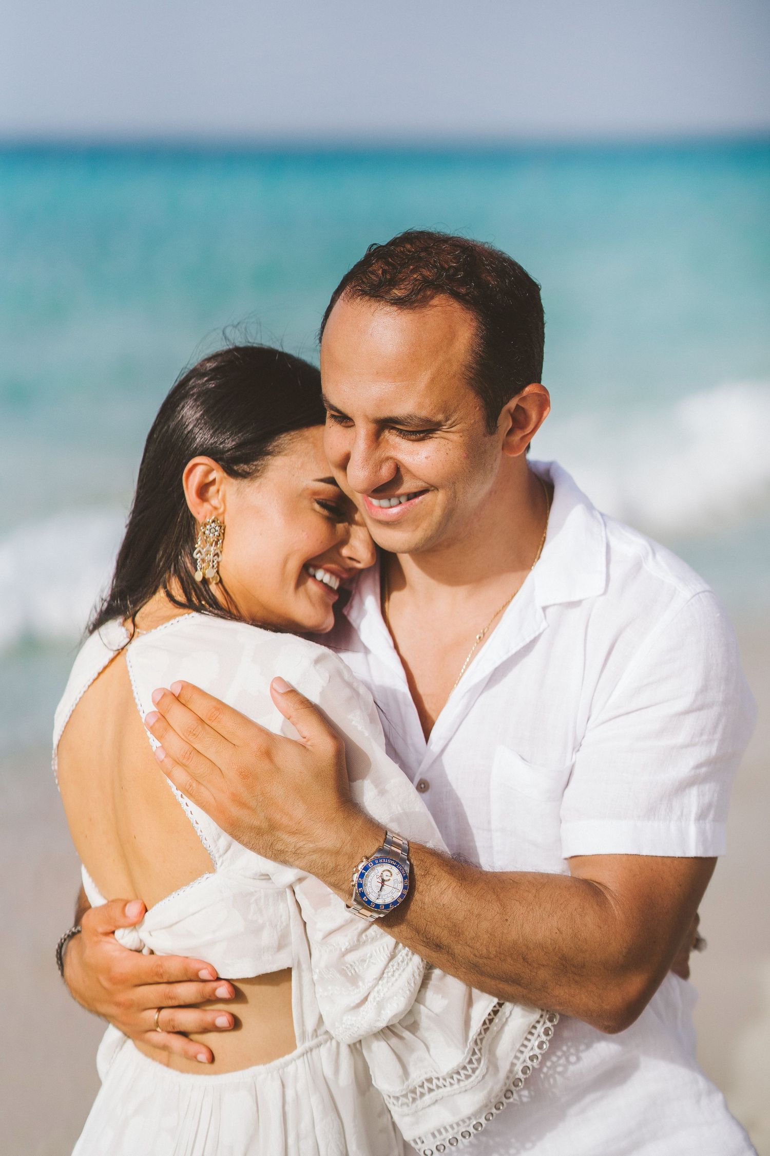 A romantic beach portrait of a couple in white embracing by turquoise waters.