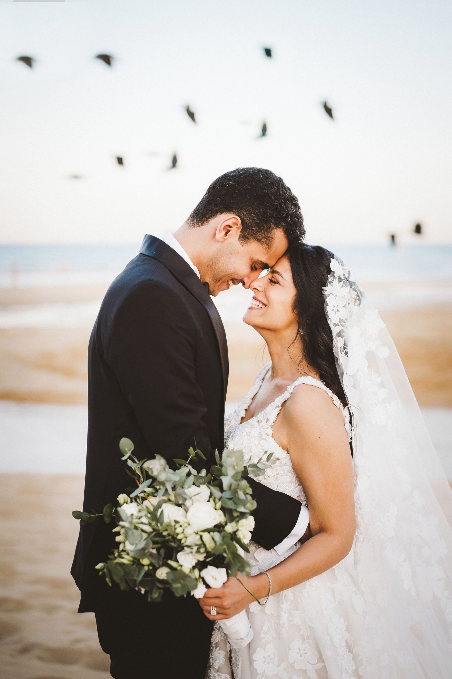 A romantic beach wedding portrait with birds flying overhead and a white floral bouquet.
