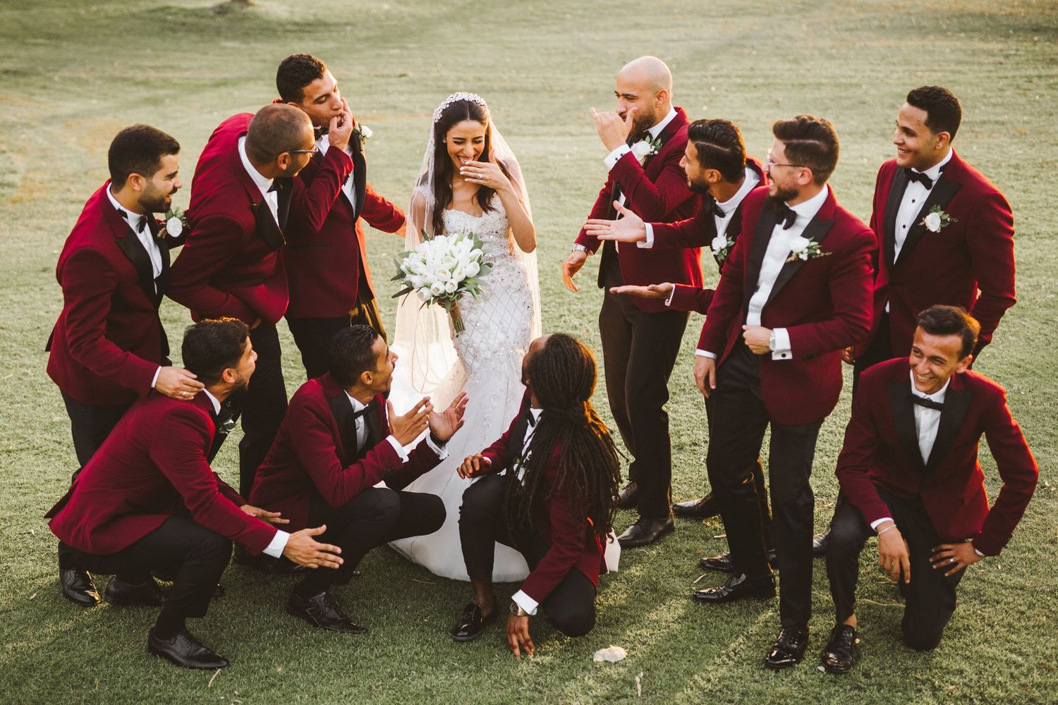 A wedding party in burgundy suits reacting playfully on a green lawn.