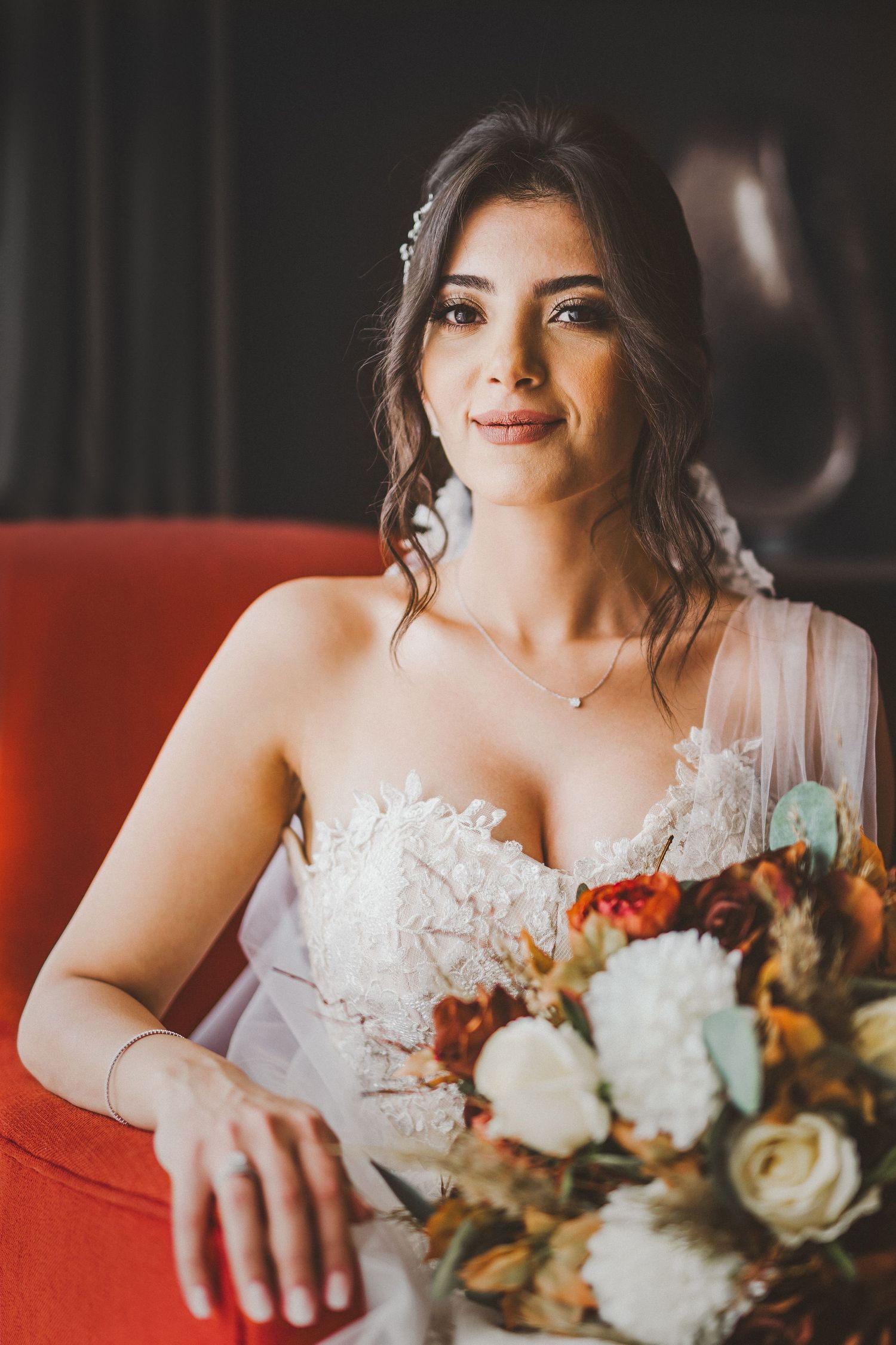 Bride in lace wedding dress sitting on red couch holding autumn-toned bouquet with roses and greenery.