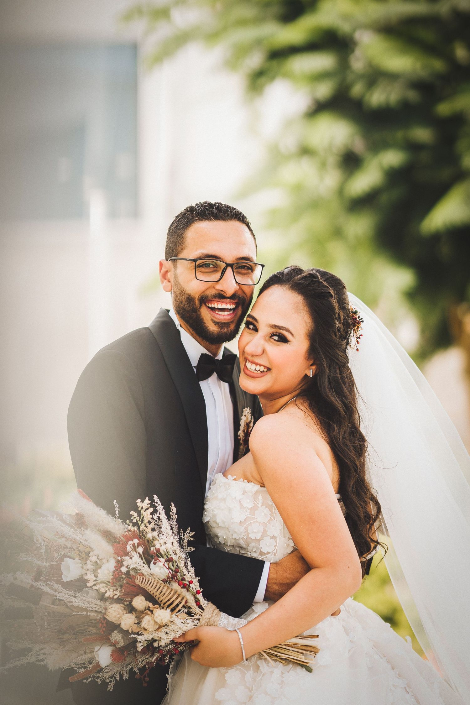 Happy newlyweds embrace and smile while holding dried flower bouquet during outdoor wedding photoshoot.