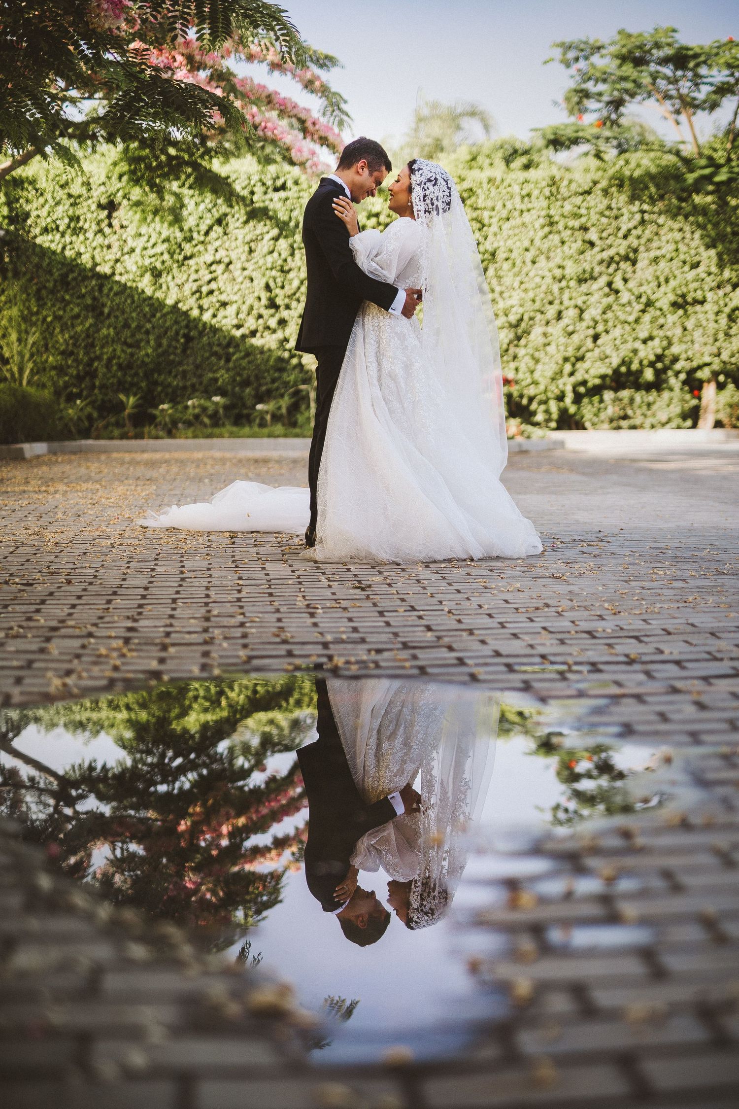 Wedding couple posing on cobblestone path with reflection in puddle beneath trees.