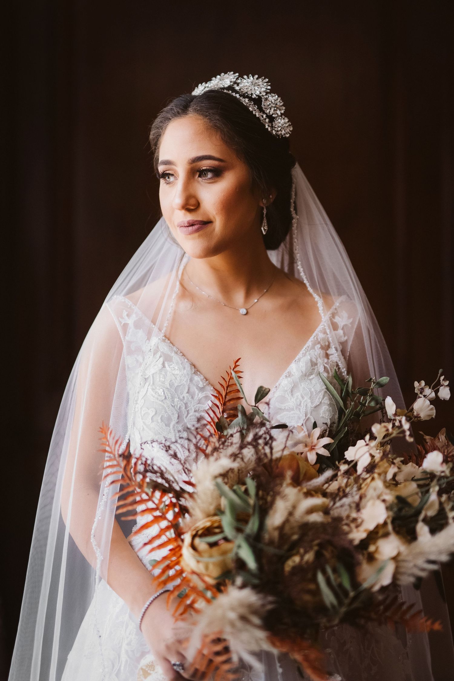 Bride in lace gown and crystal crown holds autumn-inspired wedding bouquet against dark backdrop.