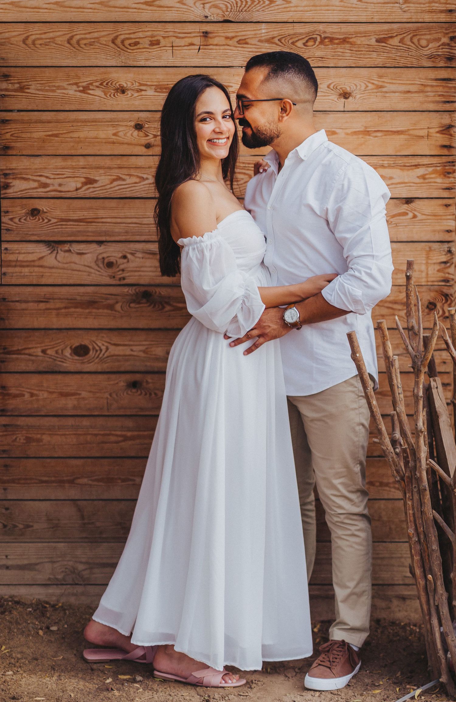 Casual engagement portrait of couple in white outfits against rustic wooden wall background.