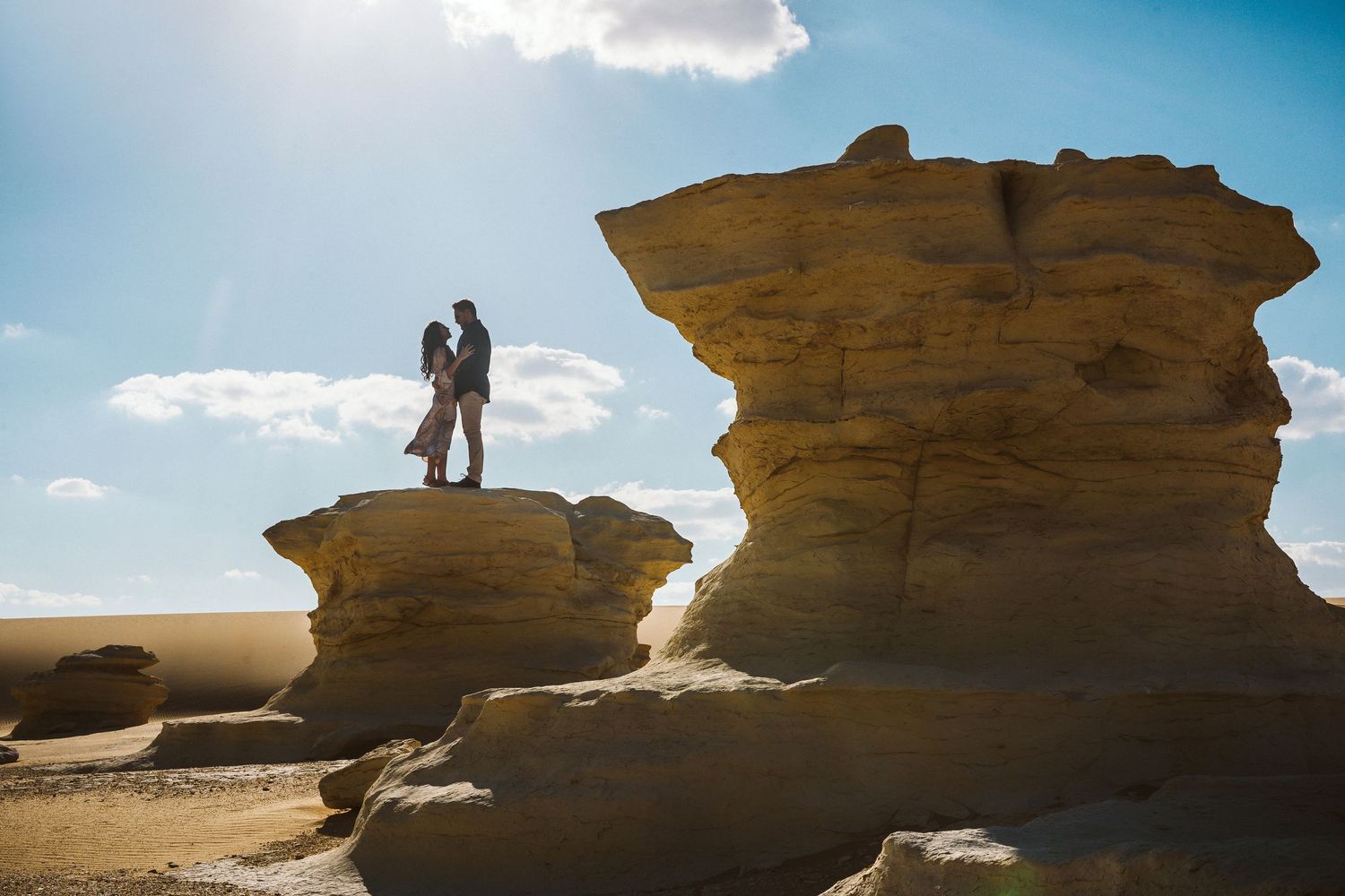 Silhouetted couple standing on rock formations against a bright blue desert sky.