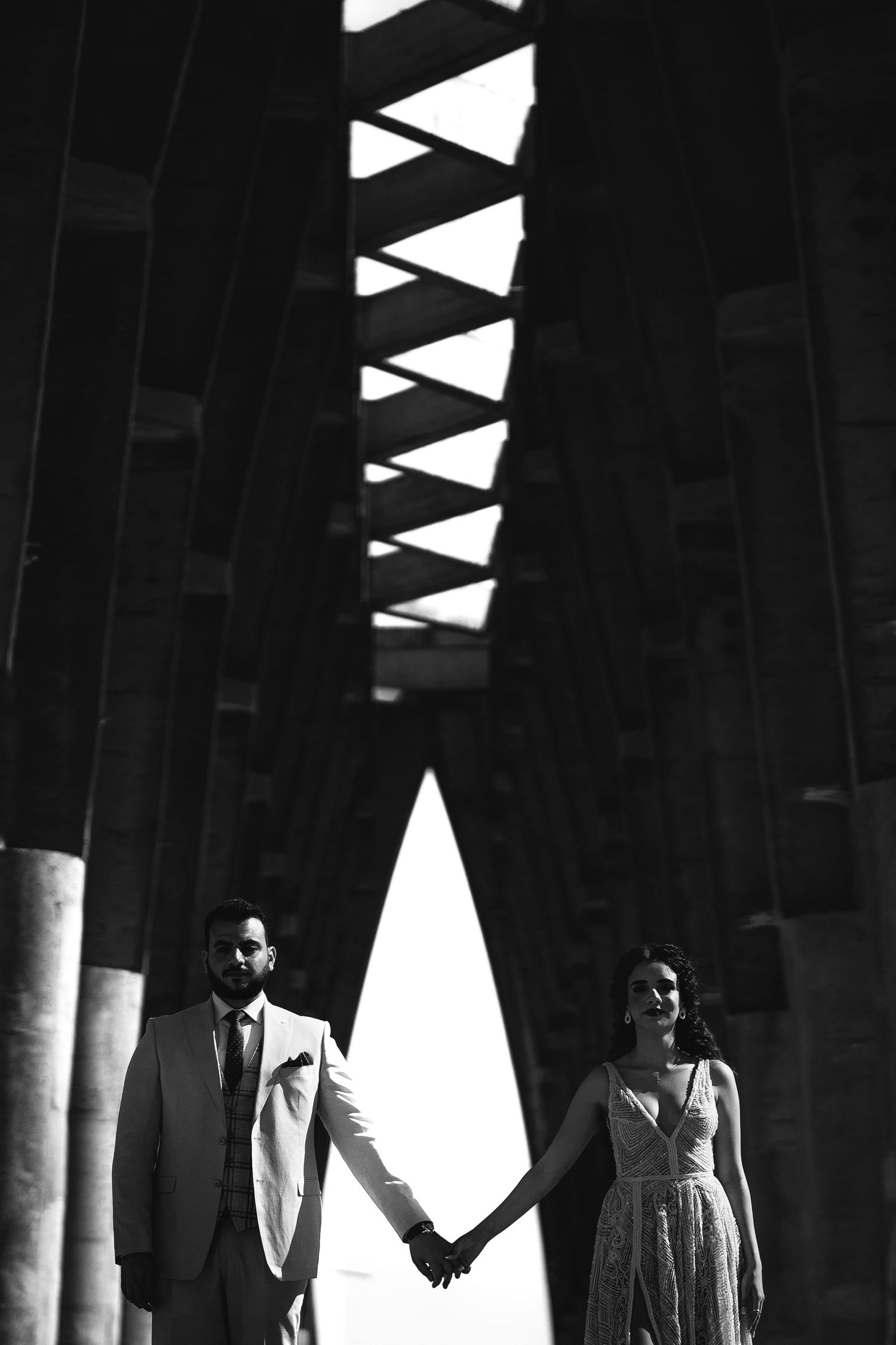 Artistic black and white photo of a couple holding hands under geometric architecture.