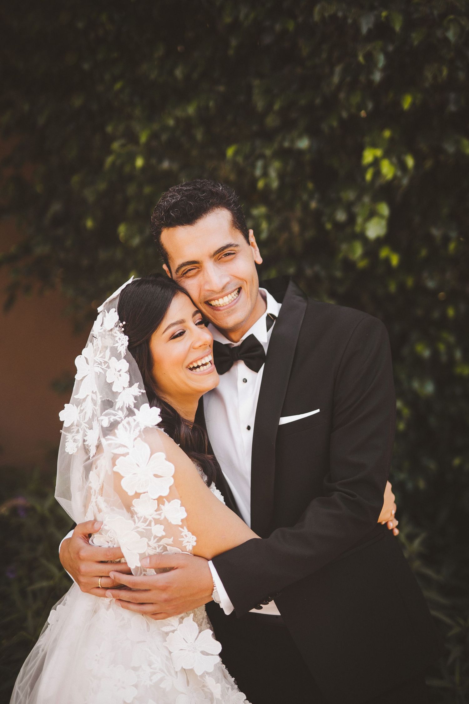 Joyful wedding couple embraces and laughs together against a green garden backdrop.