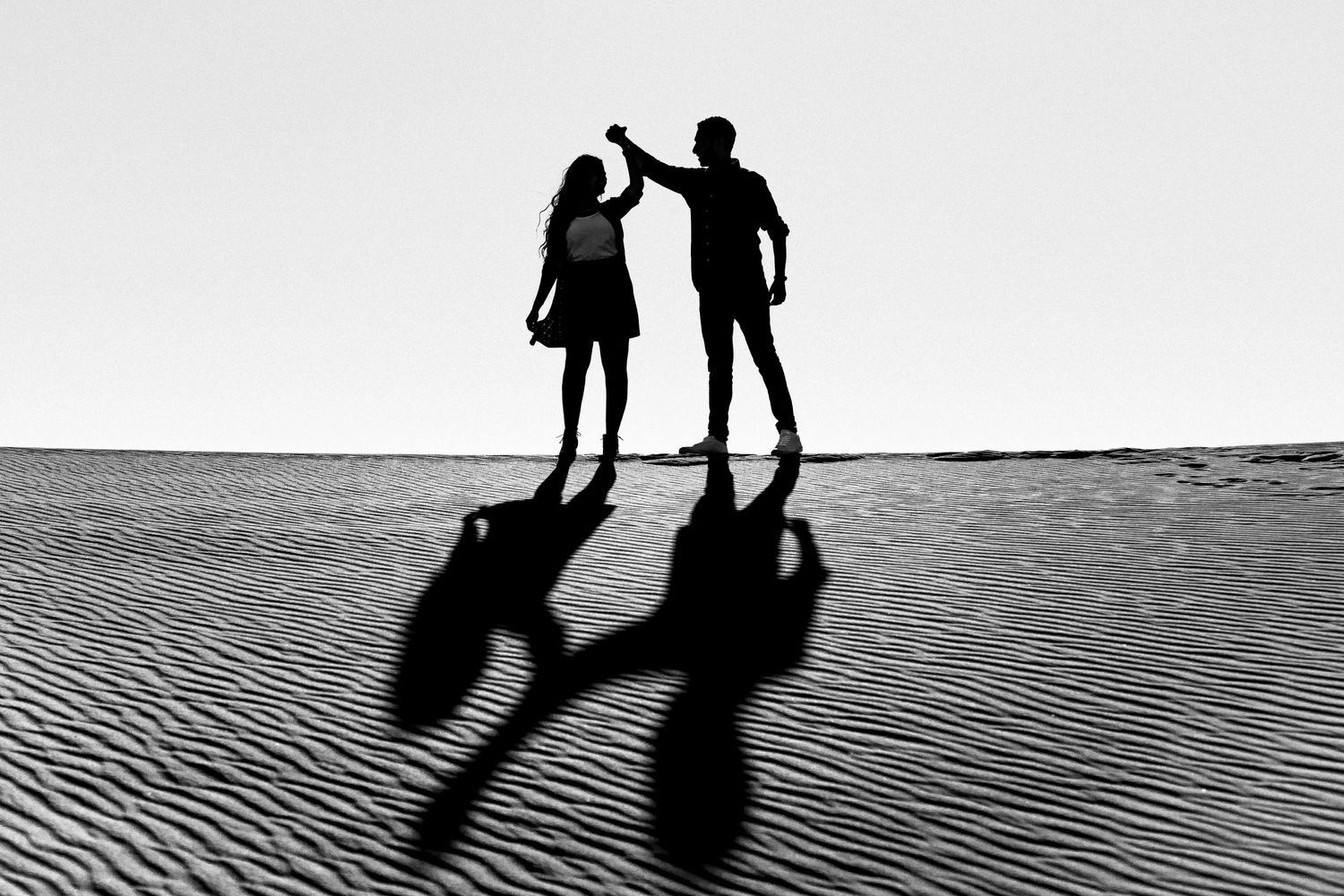 Silhouettes of two people casting long shadows on textured sand at sunset.