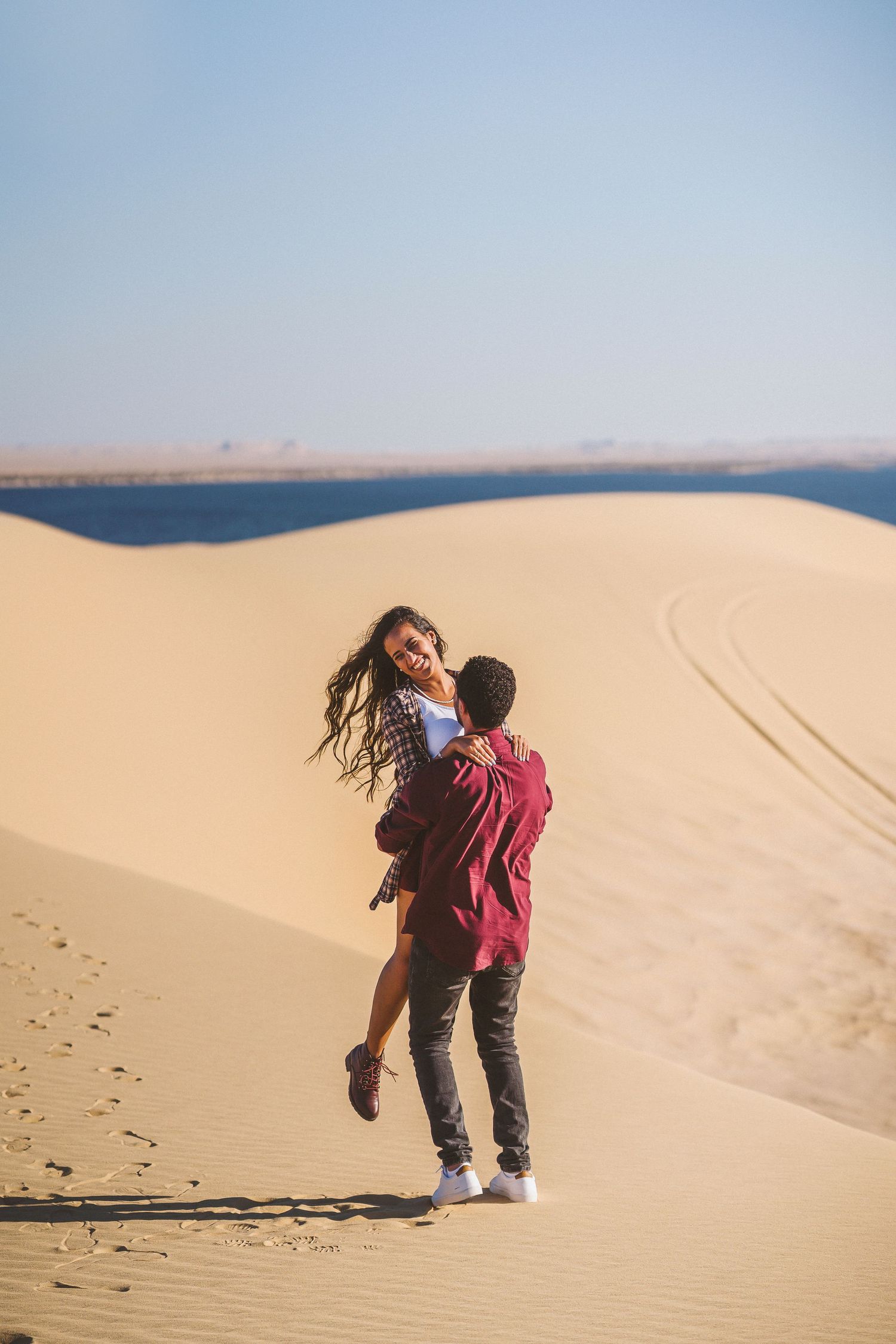 Couple playfully interacts on sandy dunes with blue sky and ocean in background.