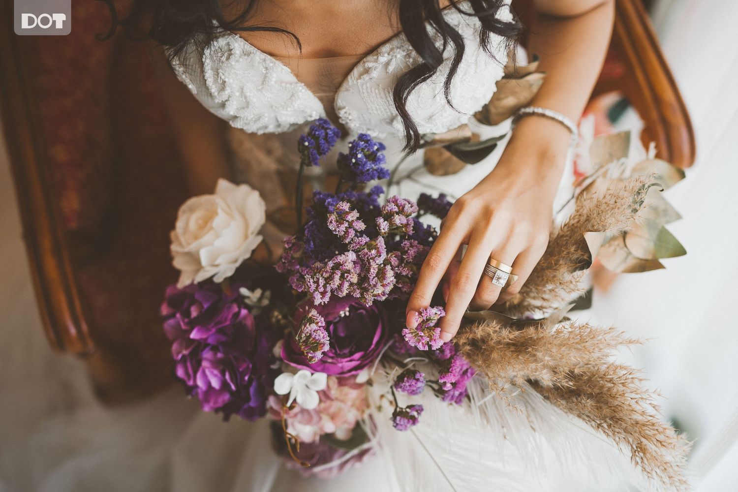 Close-up of bridal bouquet with purple flowers and pampas grass against a white wedding dress.