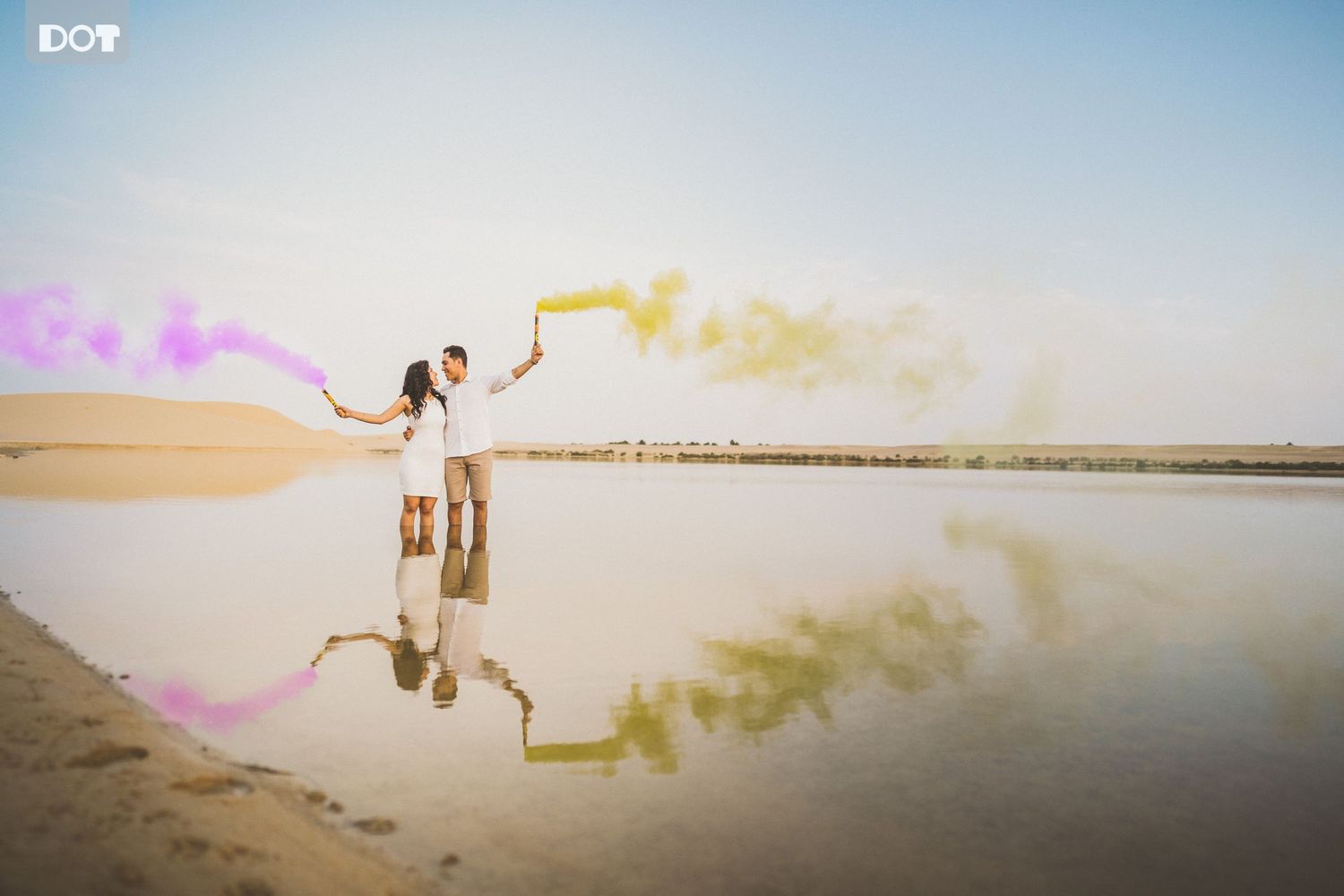 Couple poses with colorful smoke bombs at sunset by reflective water.
