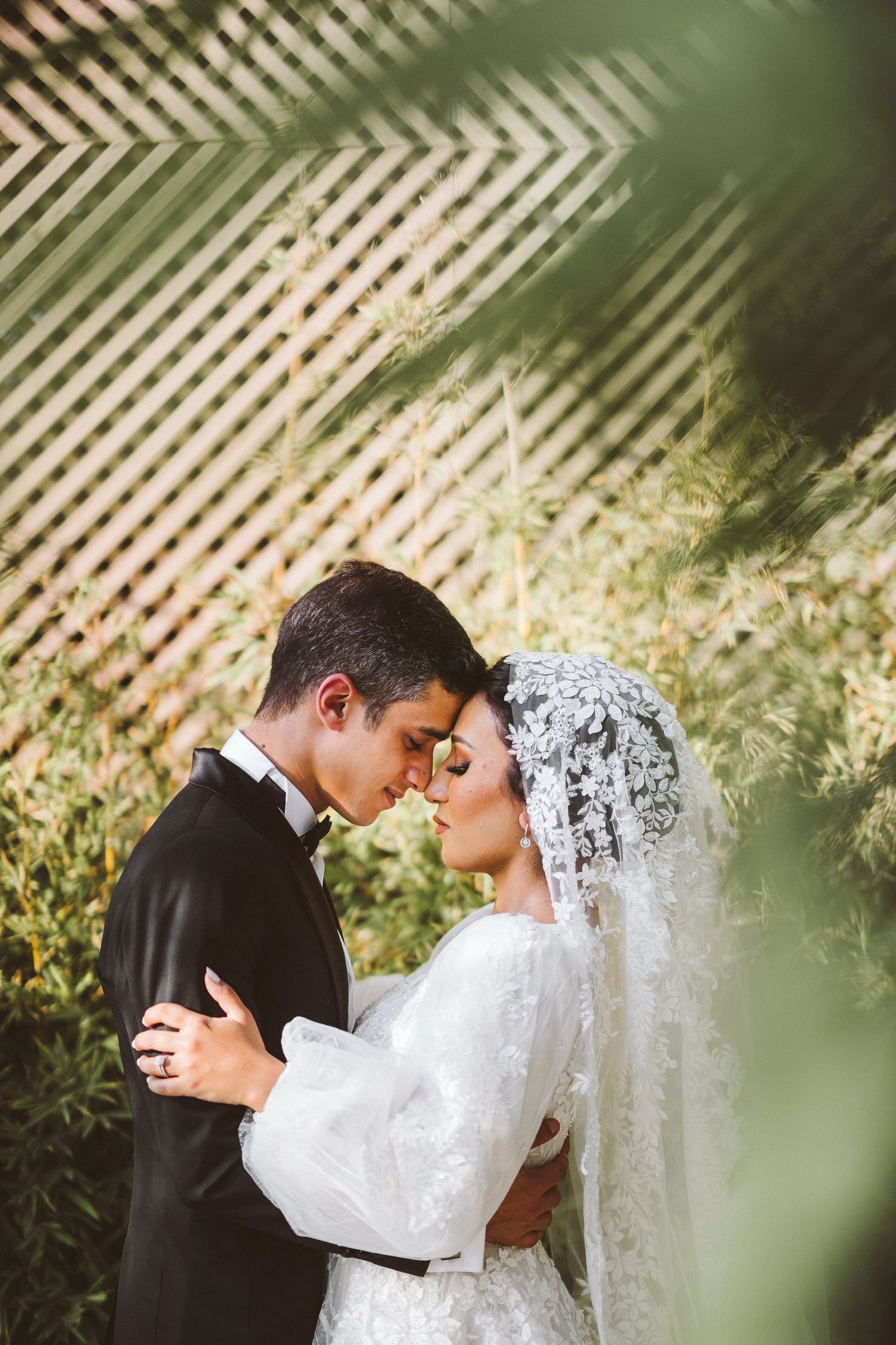 Romantic wedding portrait through foliage with bride in lace dress and traditional veil.