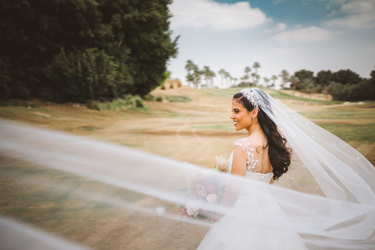 Bride with flowing veil walks along sunny path with palm trees in background.