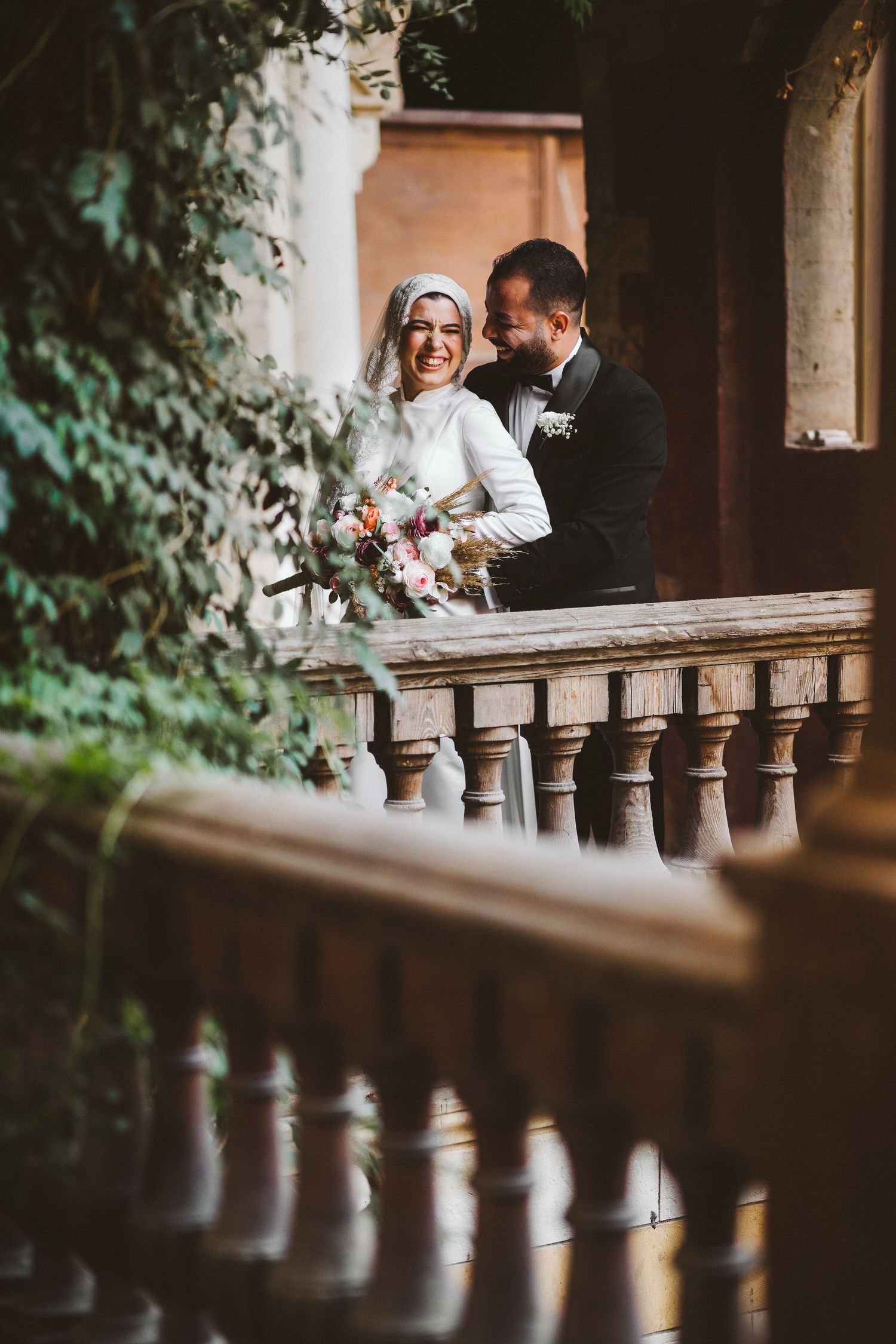 Wedding couple shares joyful moment on vintage balcony with floral bouquet.