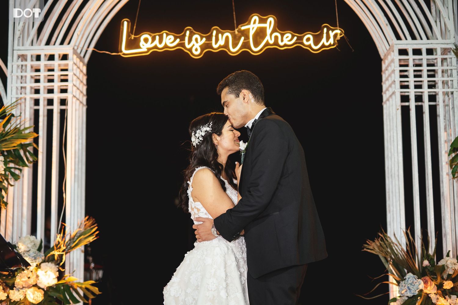 Wedding couple under illuminated 'Love is in the air' sign with floral arch.
