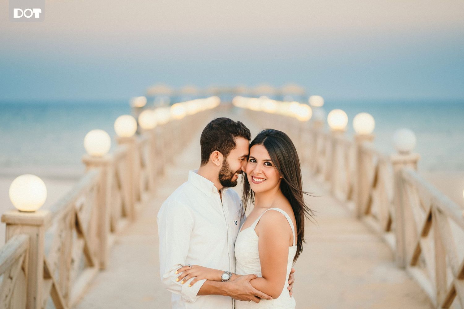 Romantic couple walks along a sunlit wooden pier over blue ocean waters.