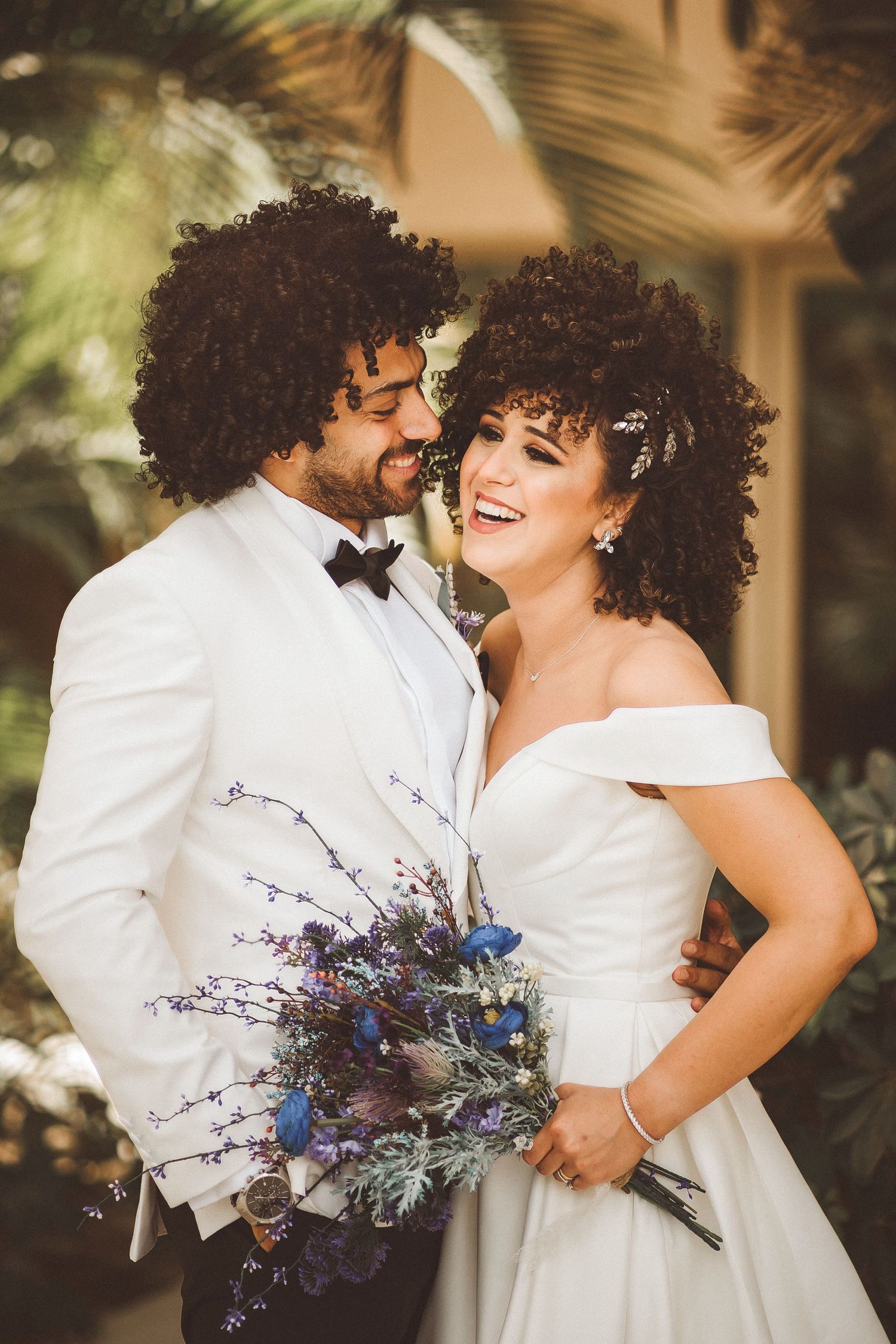 Joyful couple in white attire share a laugh while holding purple and blue floral bouquet.