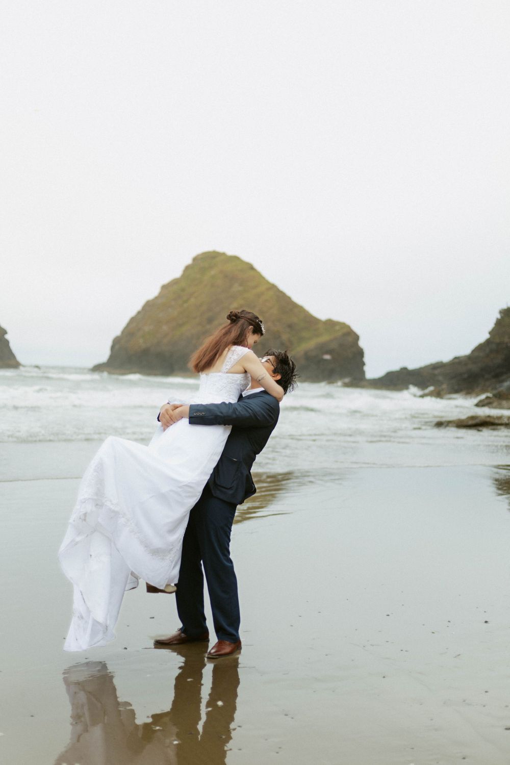 A couple in wedding attire embracing on a misty beach with dramatic coastal rock formations.