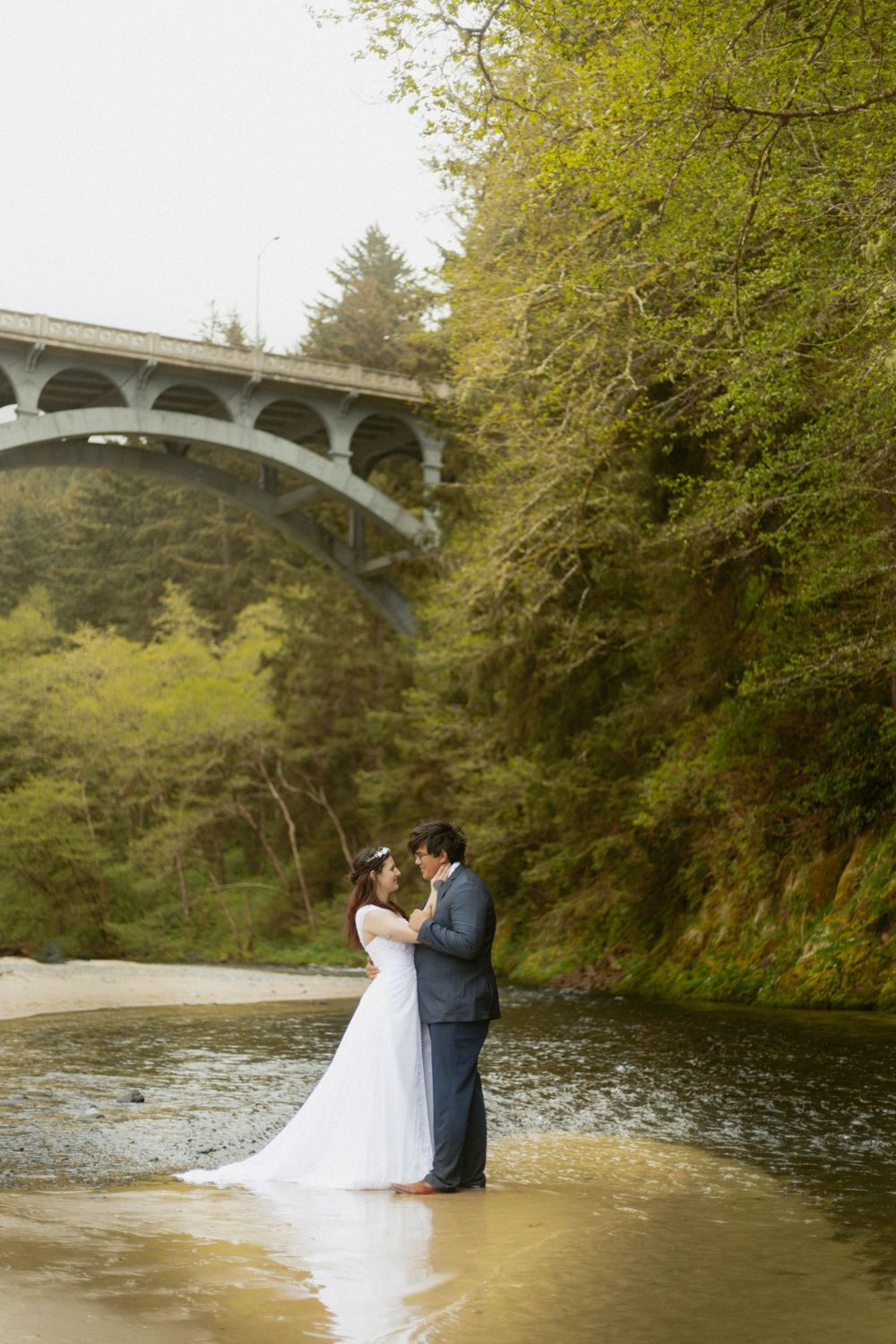 A scenic wedding photo under an arched bridge by a peaceful river surrounded by lush trees.